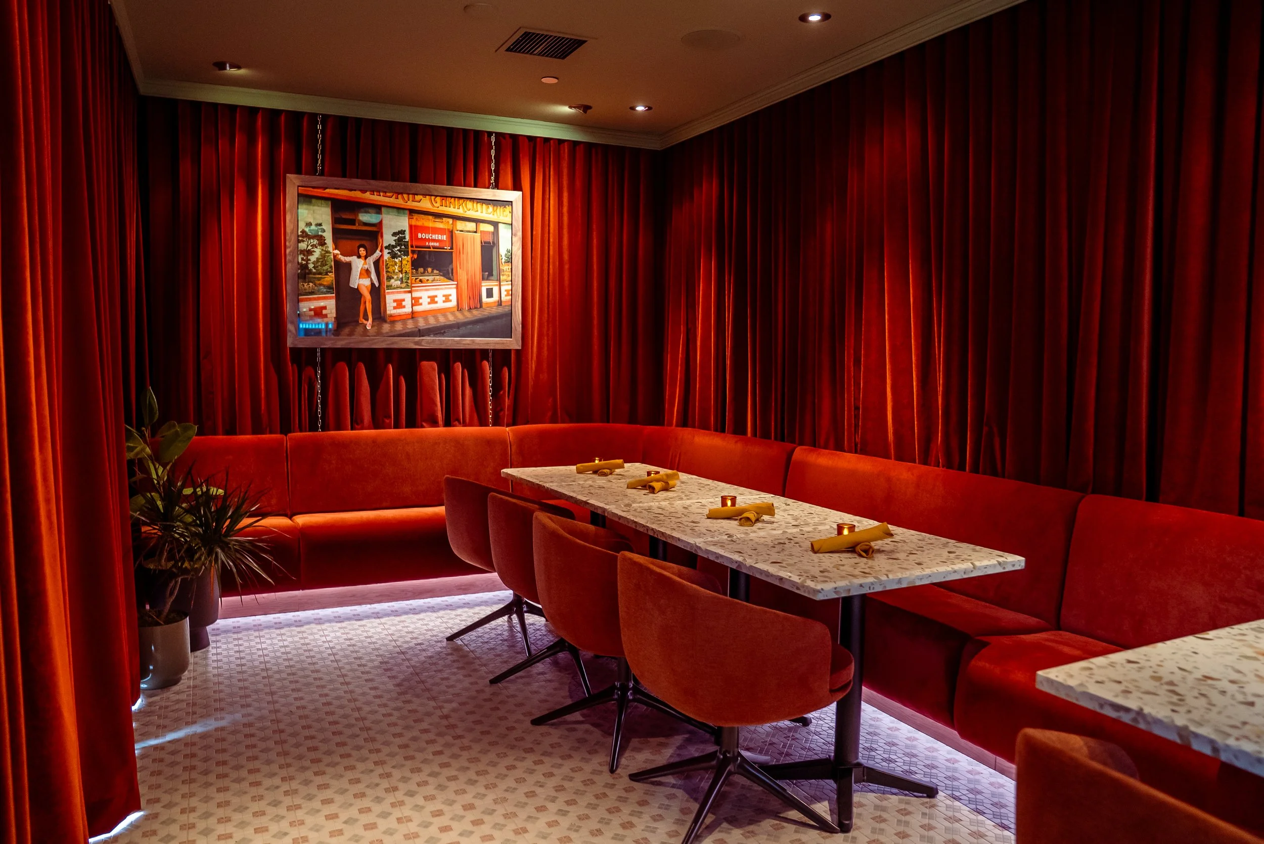 An elegantly decorated dining area with red velvet curtains, a marble table set with napkins, red velvet chairs, a corner plant, and a painting on the wall.