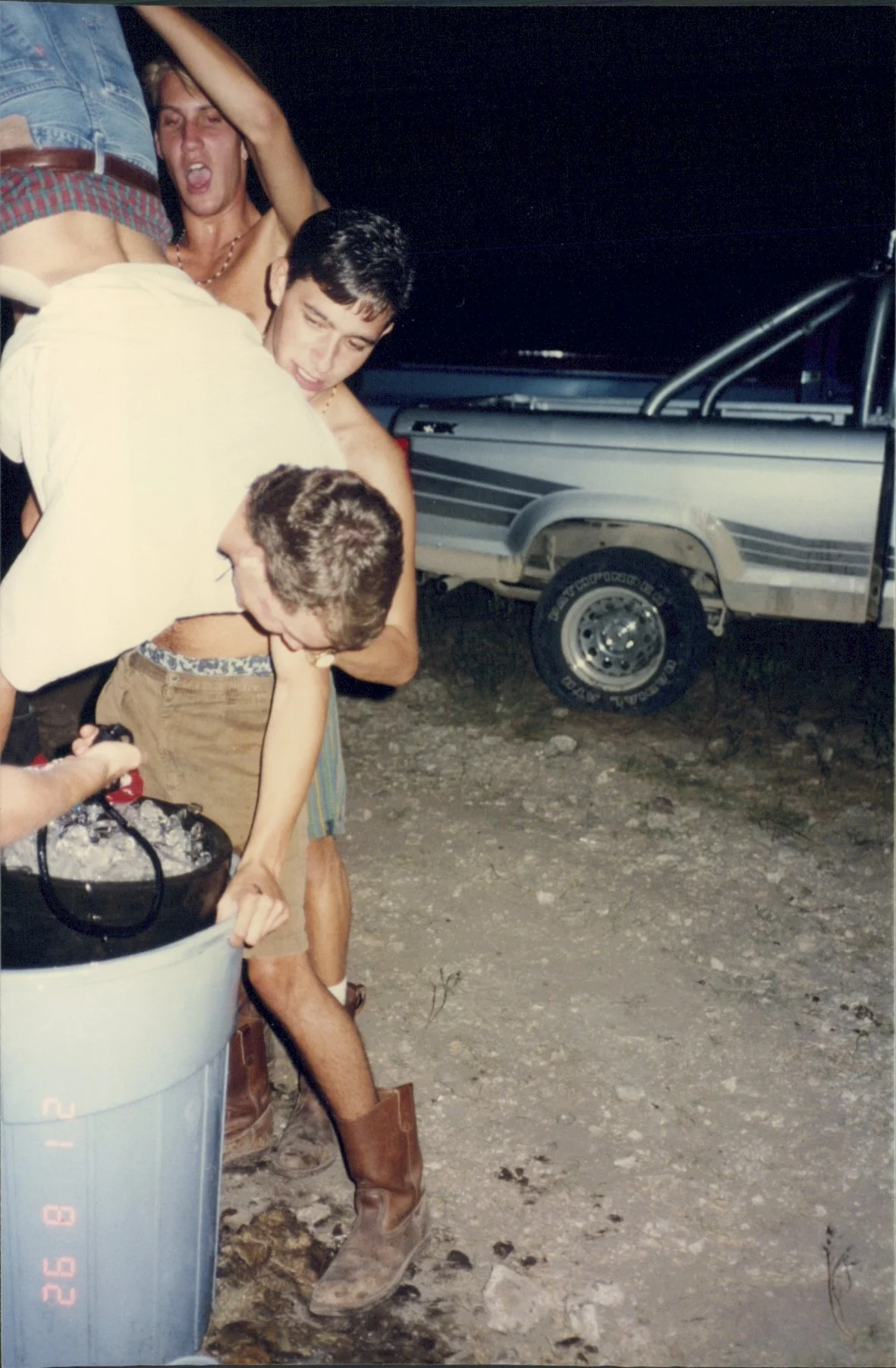 Group of young men at night, some in underwear, playing with a cooler outdoors near a pickup truck.