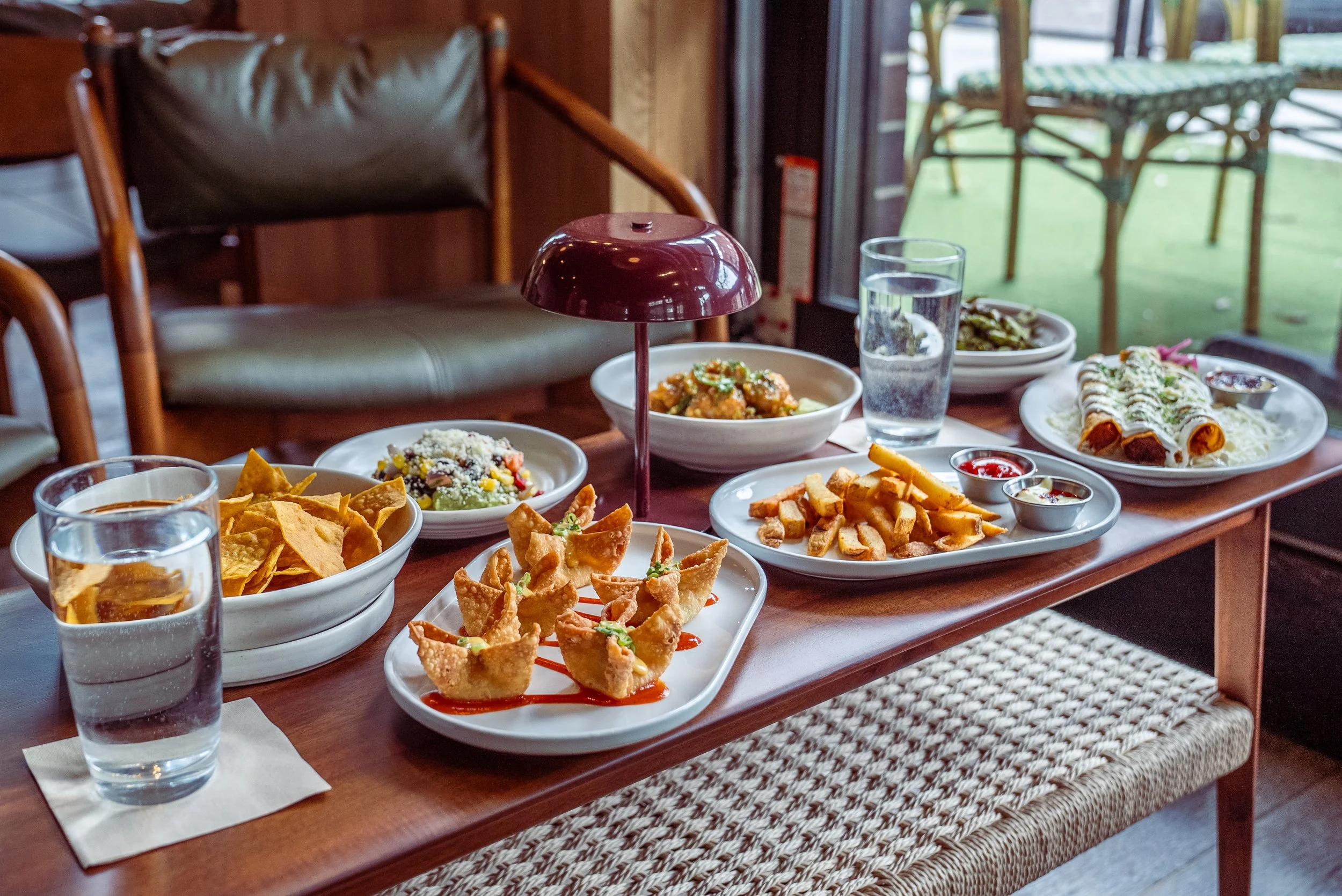 A wooden table filled with assorted Mexican dishes, including nachos, fried taquitos, a salad, small cups of salsa, and fried bread sticks, with water glasses and a red lampshade in a cozy restaurant setting.