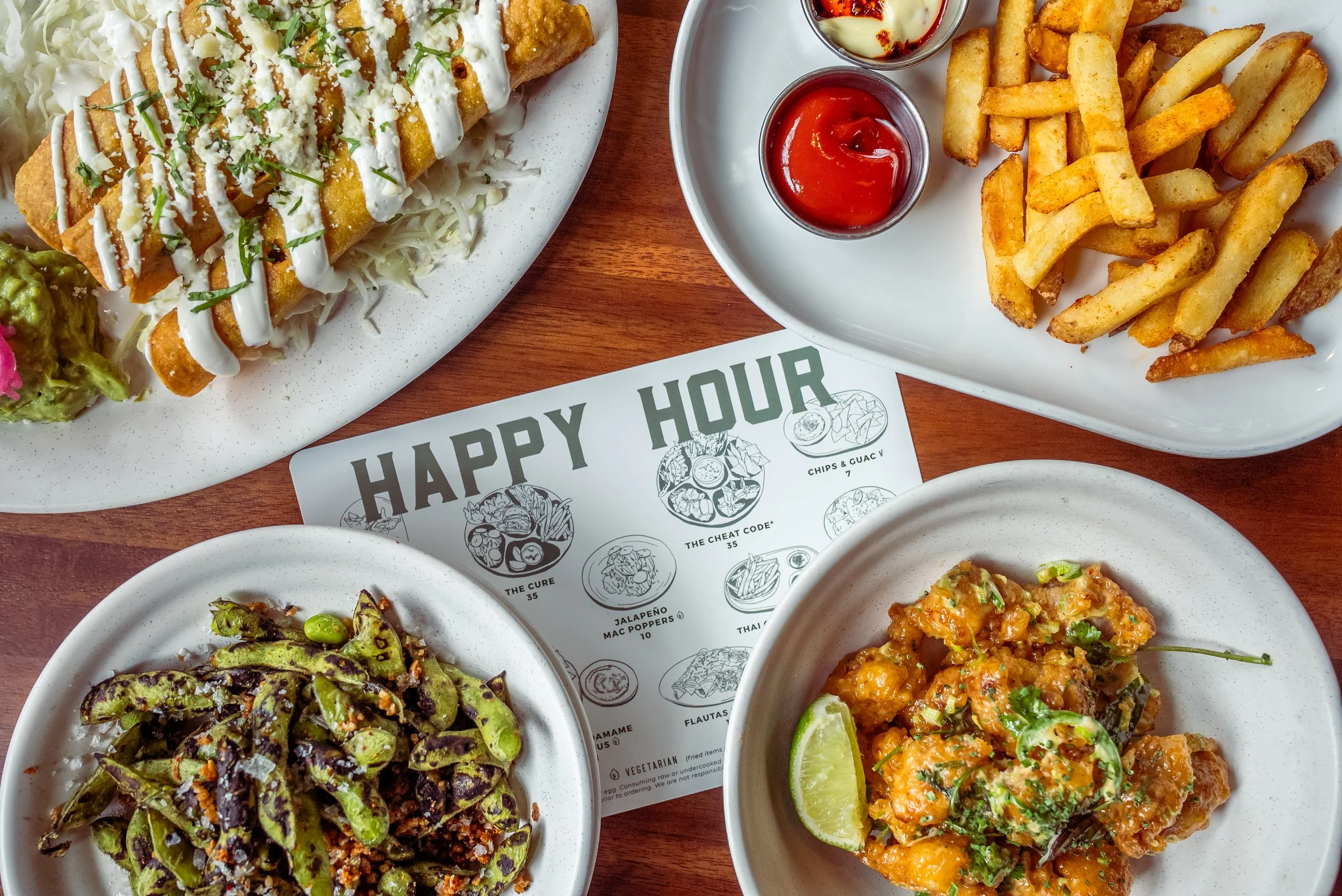 Assorted Mexican dishes on white plates, including fried fish with sauce, French fries with ketchup, roasted green beans, and spicy chicken with lime, on a wooden table with a printed menu that says 'Happy Hour'.