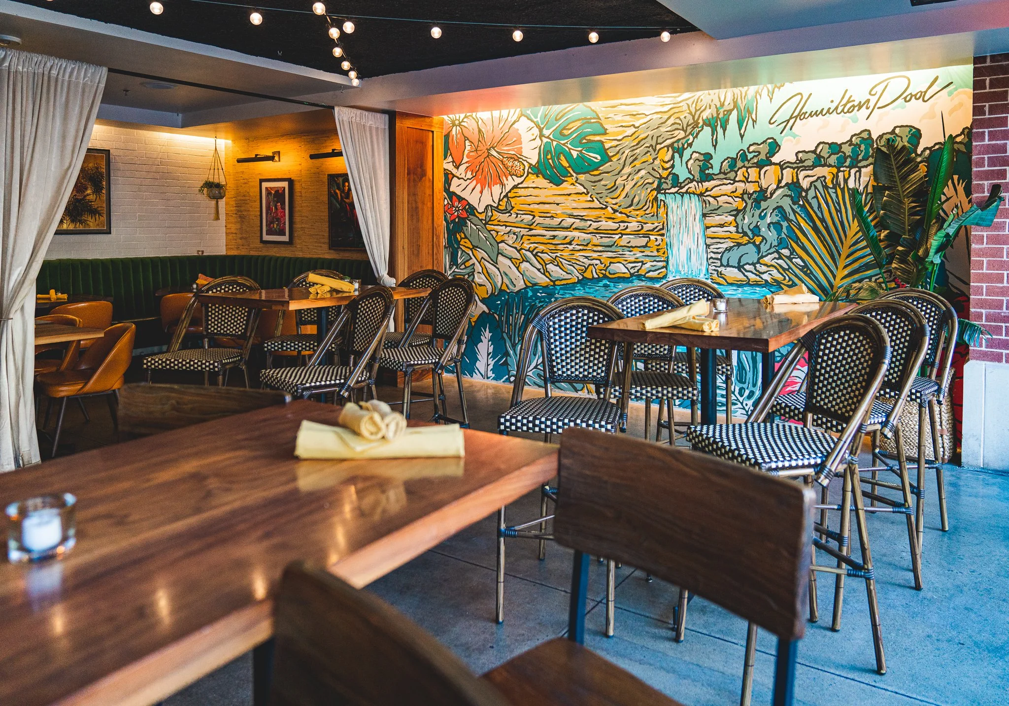 Interior of a modern restaurant with wooden tables, patterned chairs, and a vibrant mural of a waterfall and lush greenery on the wall. The ceiling has string lights and framed artwork on the brick wall.