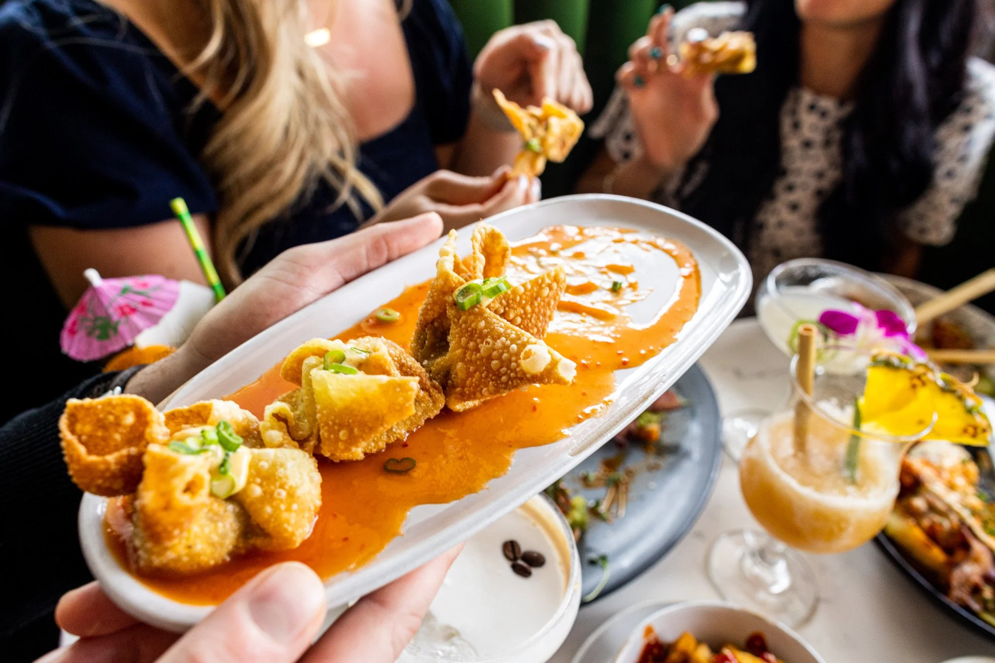 Close-up of a hand holding a white plate with fried food covered in orange sauce, served with chopped green onions, at a social gathering with people eating and drinking in the background.