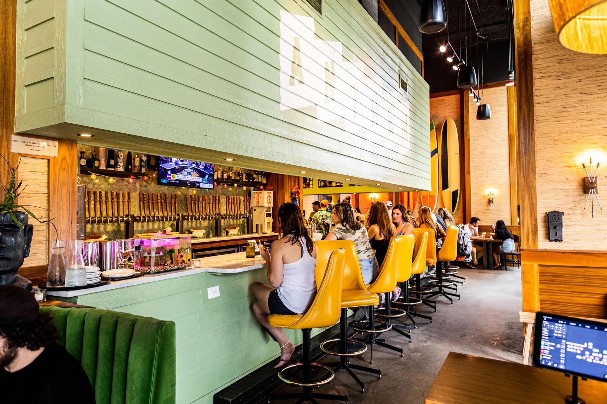Inside a lively bar or restaurant with a row of yellow bar stools and people sitting at the counter and tables, warm lighting, wood decor, and a large screen TV.