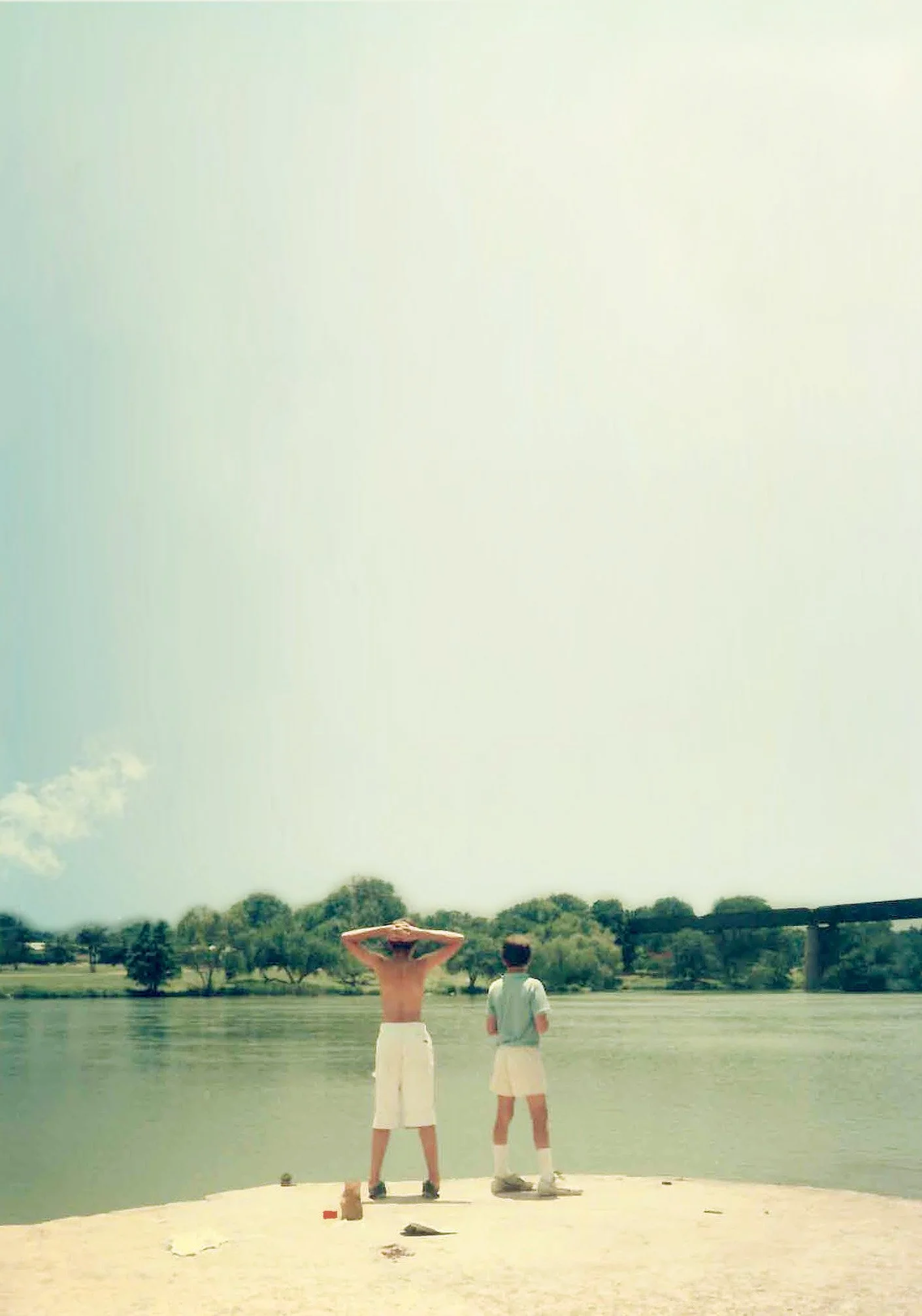 Two boys stand at a riverbank, one shirtless with hands on head, the other in a beige shirt and shorts, facing the water, with trees and a bridge in the background on a sunny day.