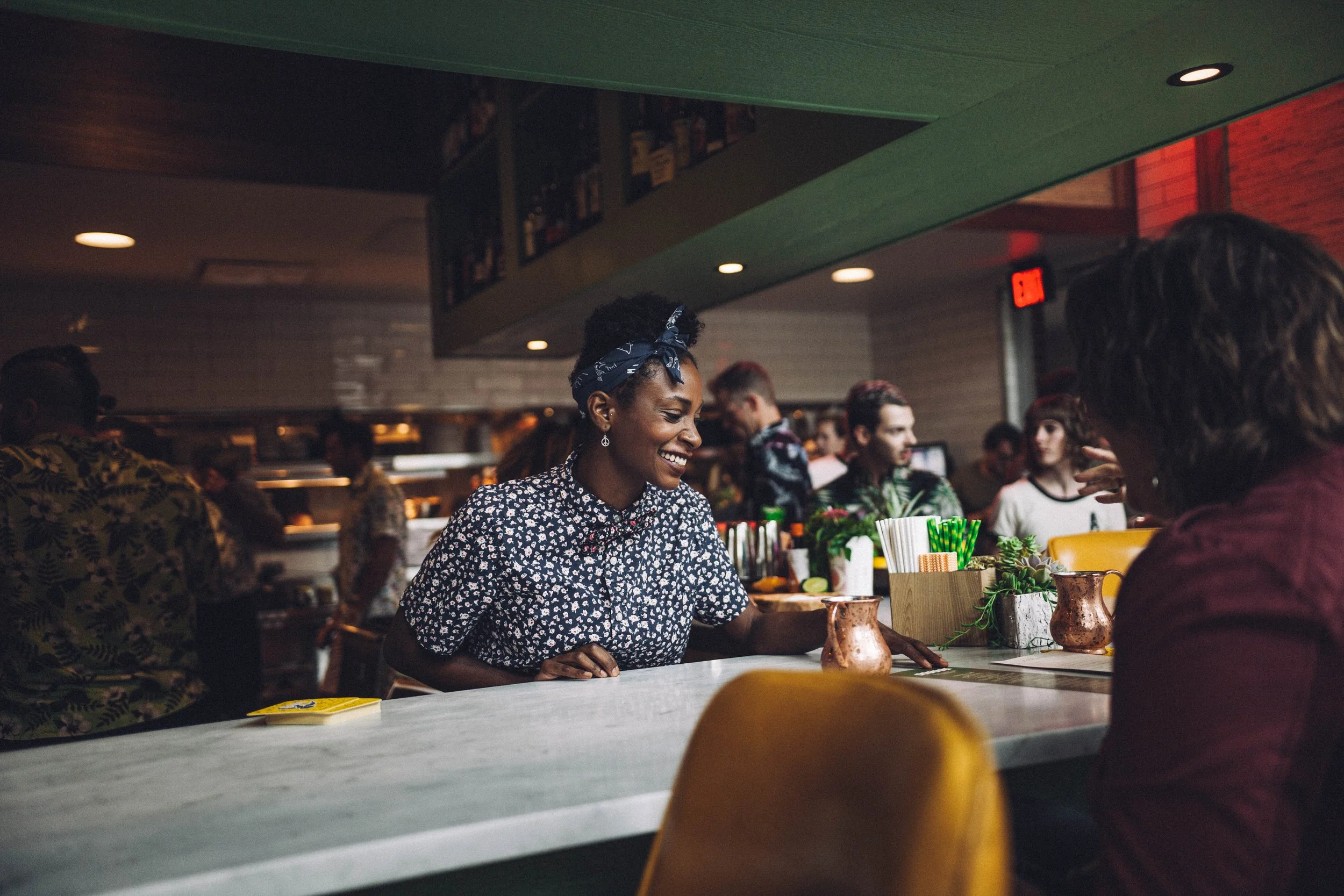 A smiling woman with a blue bandana on her head, wearing a patterned shirt, sitting at a bar counter talking to another woman in a dimly lit restaurant or bar.