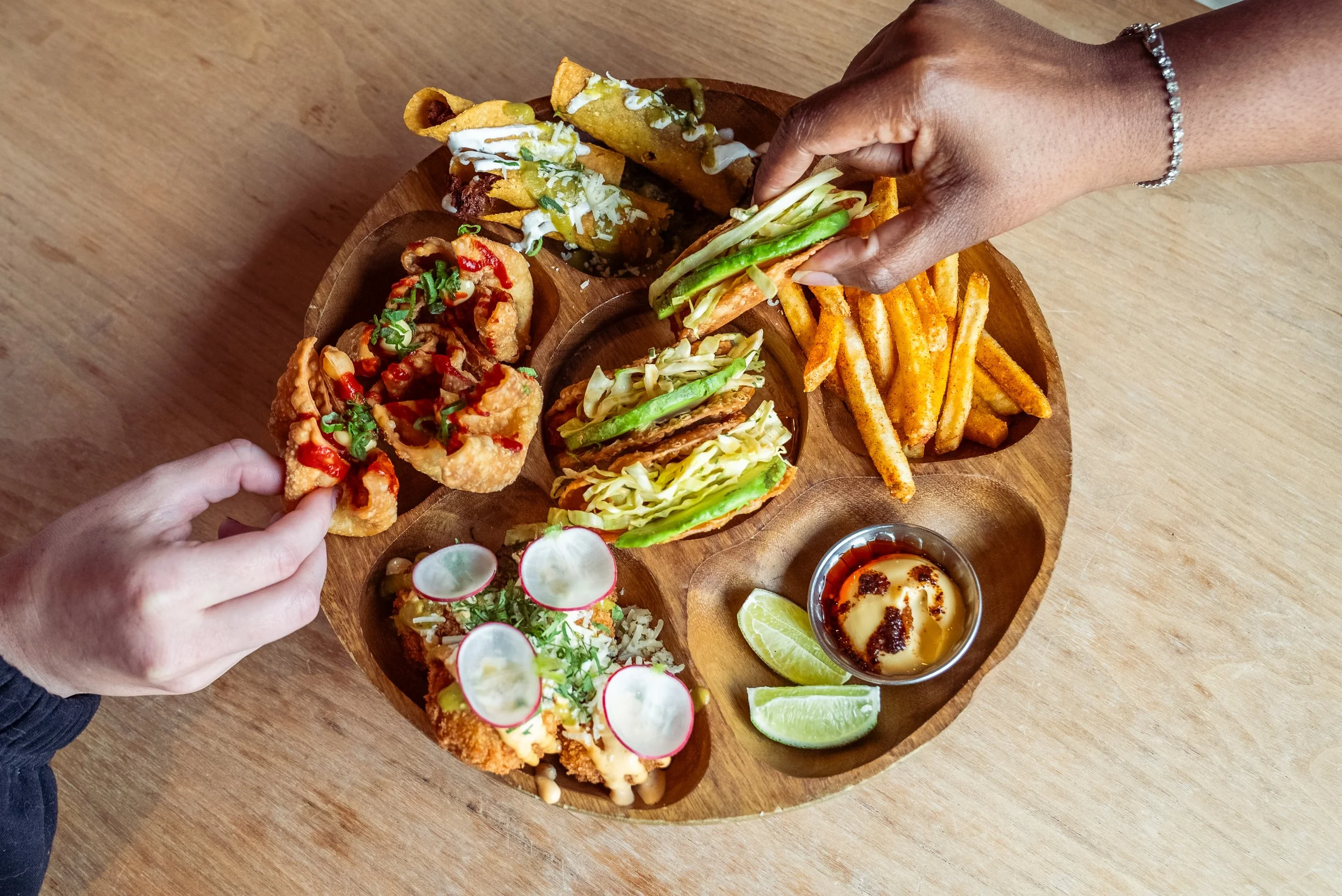 Two hands reaching for a taco on a wooden serving tray with various Mexican dishes, including fries, dipping sauce, and garnishes, on a wooden table.