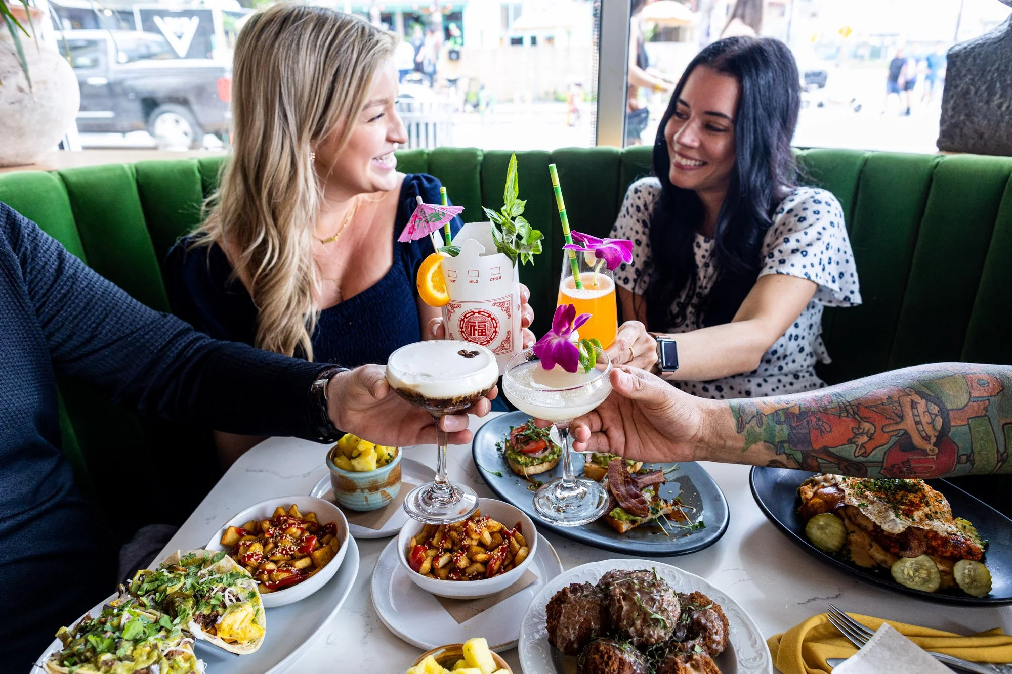Four people celebrating with cocktails and food at a restaurant table. Two women are smiling and toasting, with colorful drinks garnished with orchids and umbrella decorations. The table has various dishes including salads, appetizers, and main courses, with a green booth seating and large windows in the background.