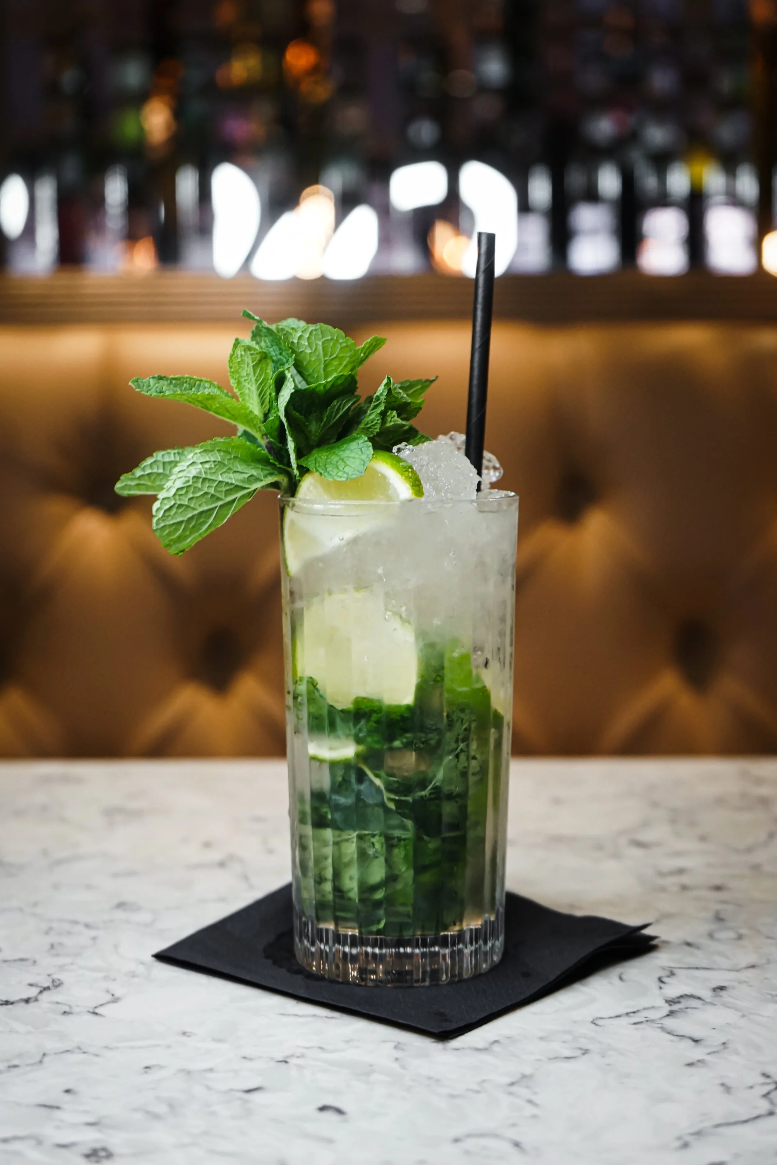 A tall glass of mojito cocktail with lime wedges, fresh mint leaves, crushed ice, and a black straw, placed on a black napkin on a marble table.