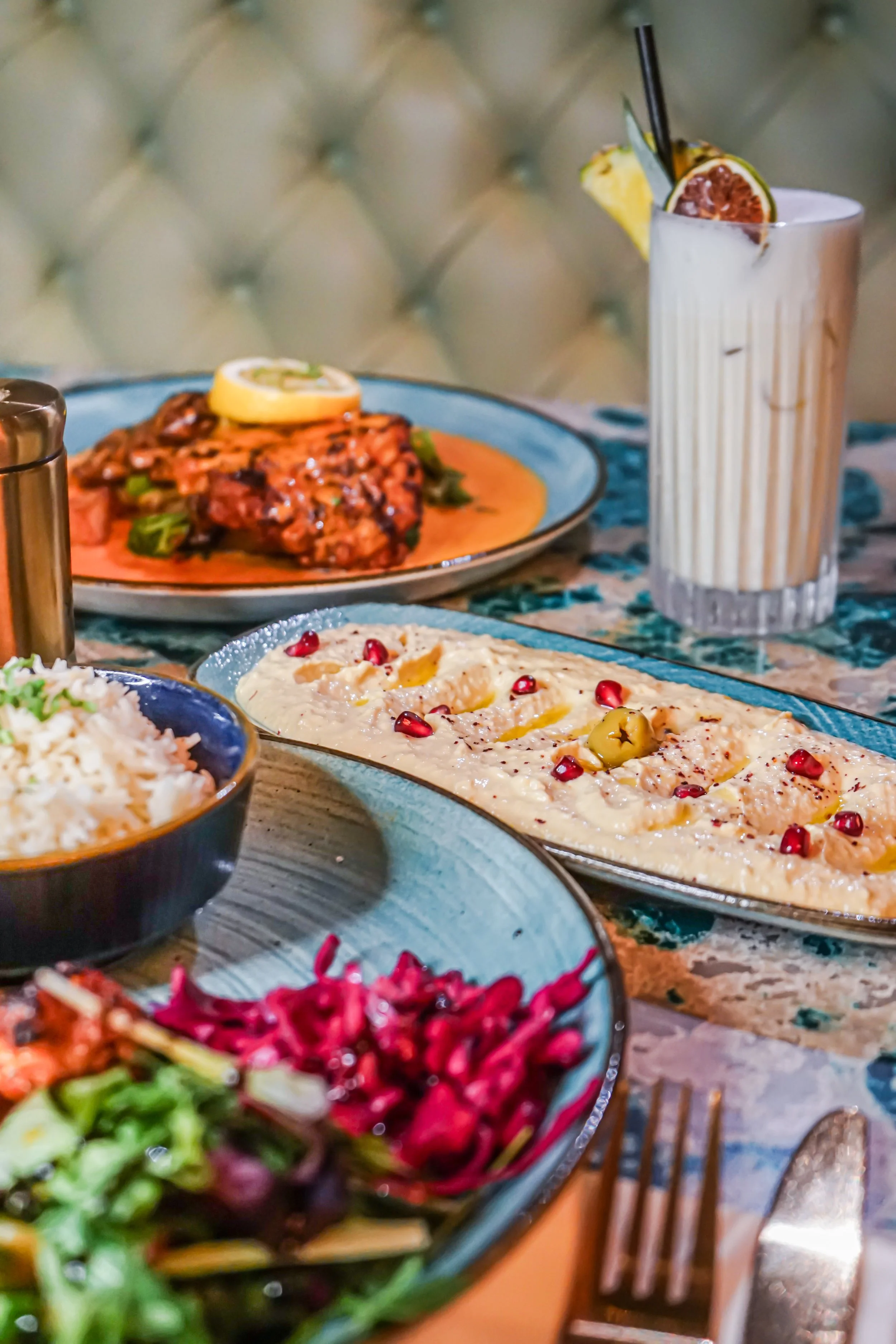 A table set with various dishes including a creamy dish garnished with pomegranate seeds, a salad with greens and vegetables, a plate with a spicy meat dish topped with lemon slices, and a tall glass of a creamy beverage garnished with a passion fruit wedge and a black straw.