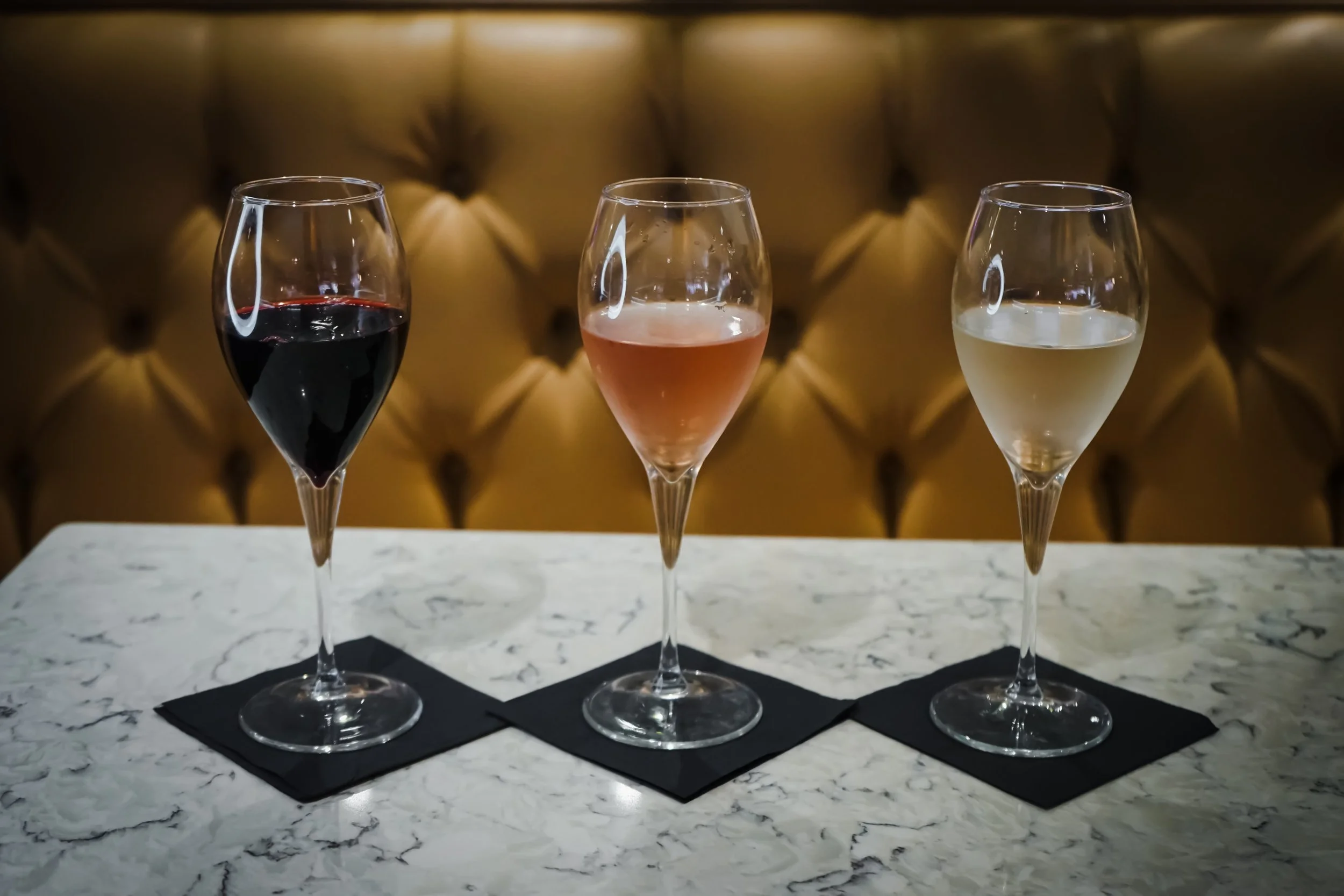 Three wine glasses on a white marble table, each filled with different types of wine: red, rosé, and white, with a tufted leather background.