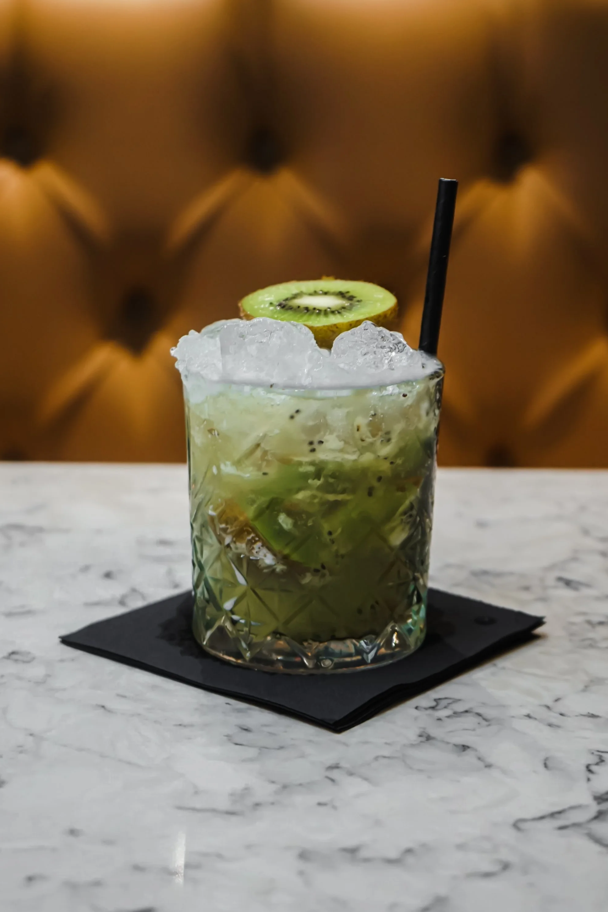 A glass filled with a green kiwi cocktail topped with crushed ice, garnished with a slice of kiwi, with a black straw, placed on a black napkin on a white marble surface.