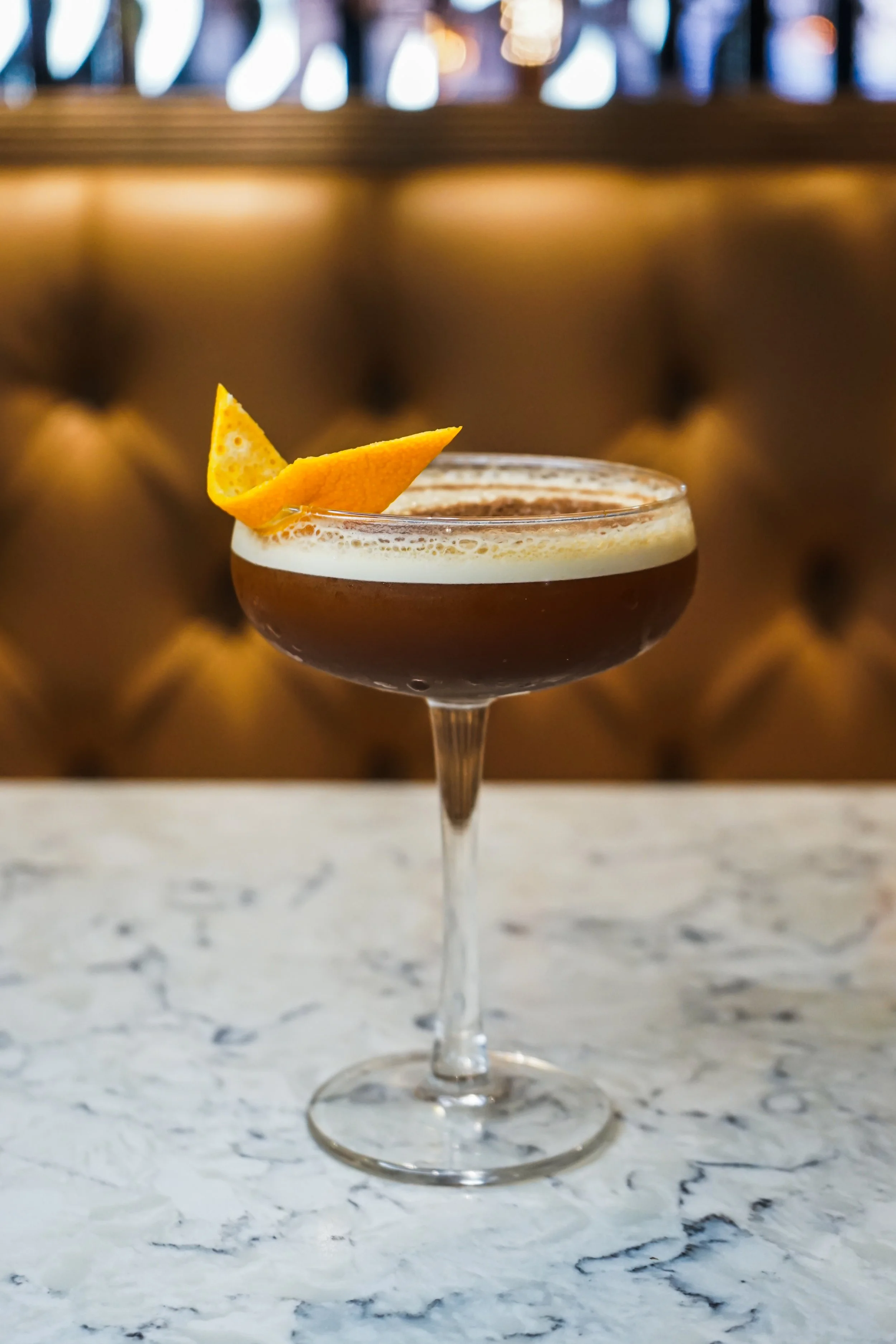 A cocktail glass sitting on a white marble surface with an orange twist garnish. The background is a blurred tufted leather seating with warm lighting.