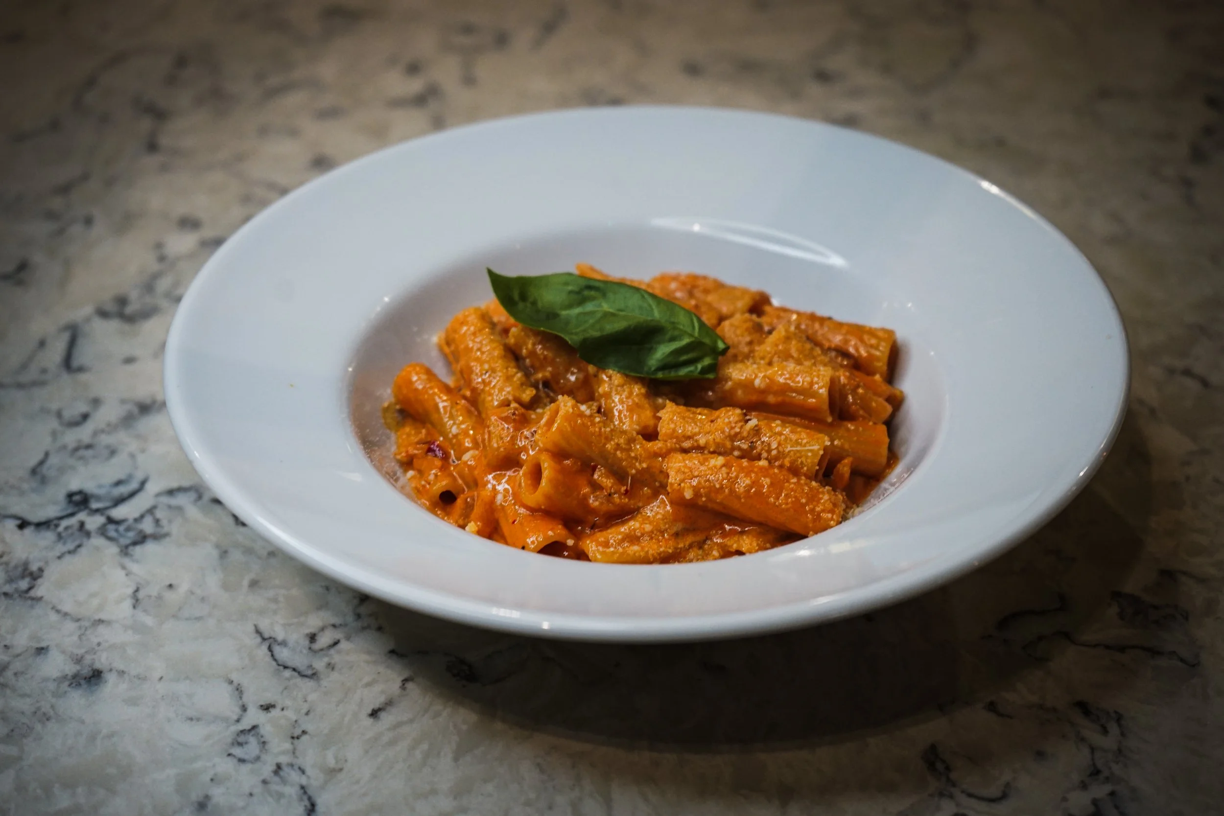 A white bowl with cooked penne pasta in a tomato sauce, garnished with a fresh basil leaf, on a marble countertop.