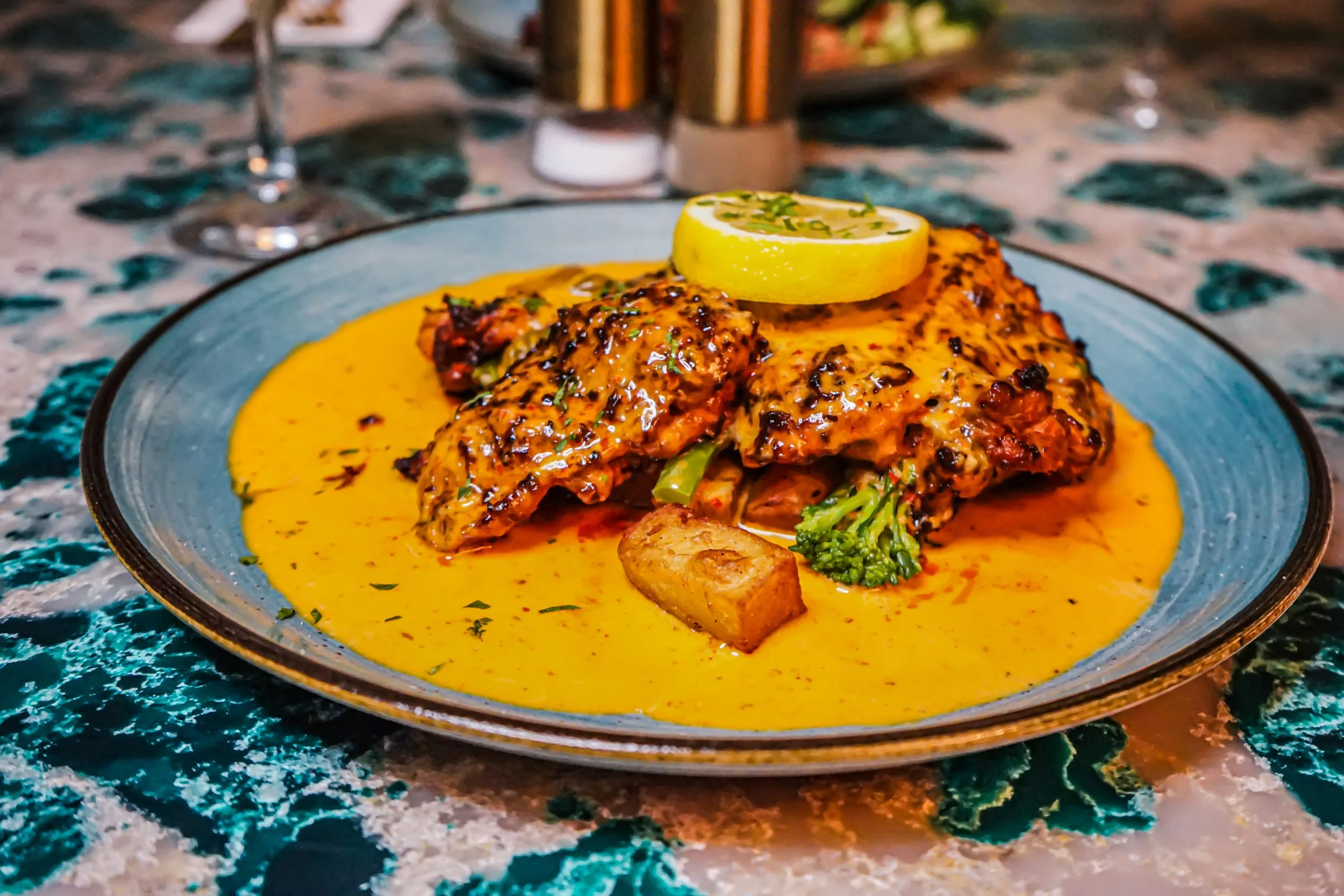 A plate of Indian curry with a lemon slice garnish, accompanied by a piece of bread, on a vibrant, patterned table.