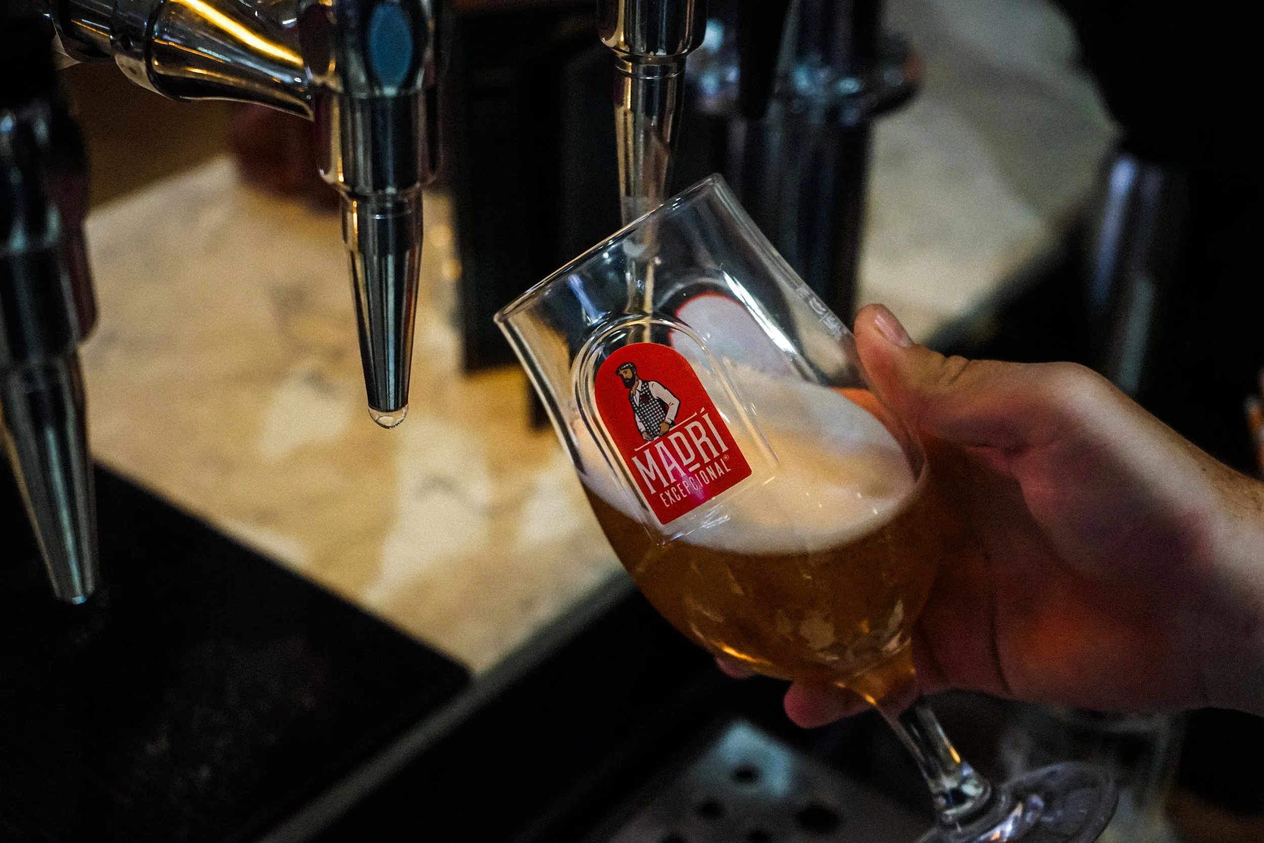 A hand holding a glass of beer with a logo that says 'Madri' underneath a cartoon image of a man. The glass is being filled from a beer tap.