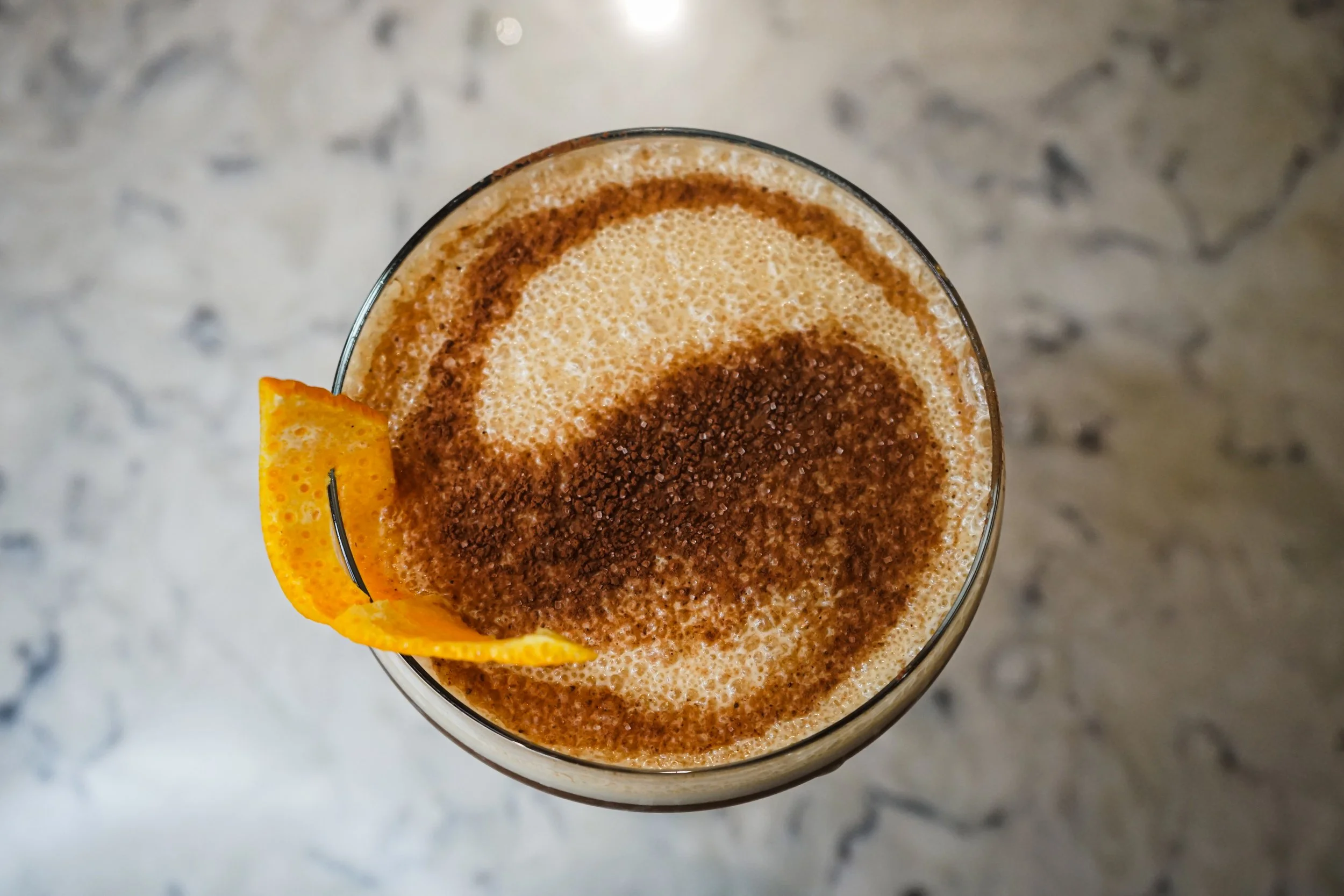 Close-up of a drink topped with frothy foam, garnished with a twist of orange peel, on a speckled gray marble surface.