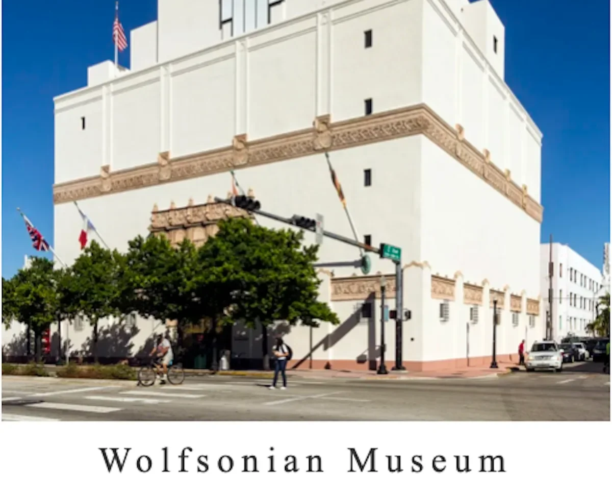 Exterior view of the Wolfsonian Museum, a white historic building with decorative architectural details, located on a city street corner with pedestrians, bikes, and cars, under a blue sky.