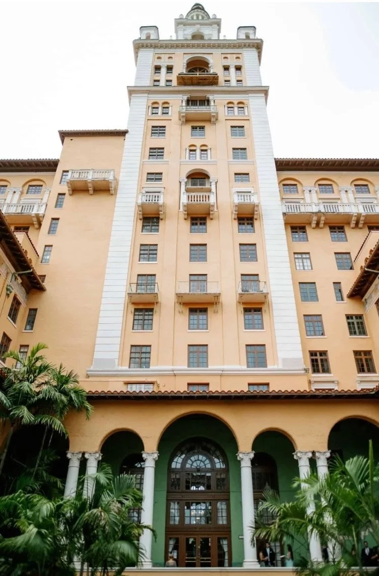 A tall, historic-looking building with a beige facade, white accents, and multiple small windows. The entrance features large green archways with white columns, surrounded by lush greenery.