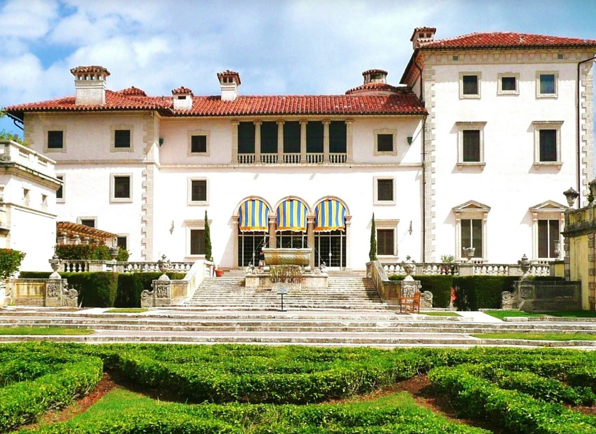 A large white mansion with multiple floors and red tiled roofs, surrounded by a manicured garden with steps leading up to the entrance and a fountain in front.