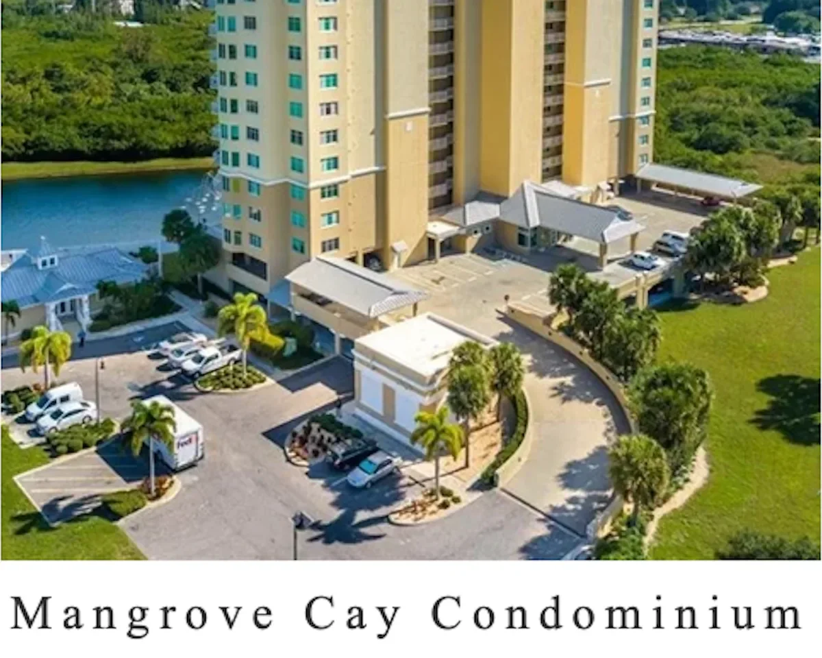An aerial view of Mangrove Cay Condominium, a high-rise apartment building with parking lots, greenery, palm trees, a pond, and landscaped areas surrounding it.