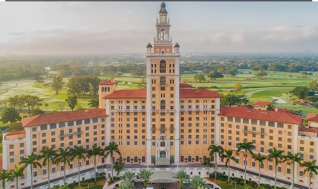 An aerial view of a large luxury hotel with a central tower, surrounded by green golf course and trees, under a partly cloudy sky.