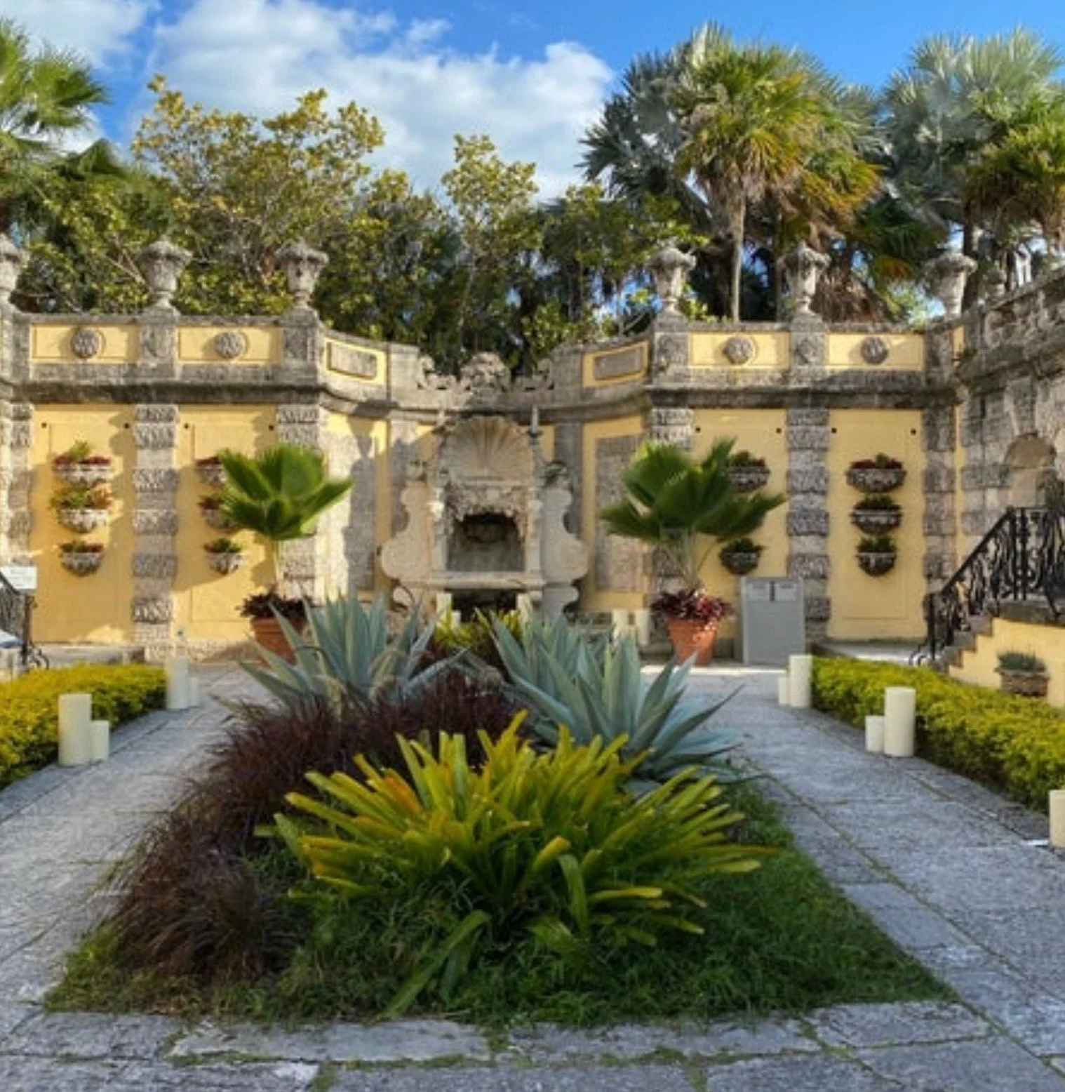 A historic yellow and gray stone building with ornate architectural features, surrounded by well-maintained gardens and tropical plants, under a partly cloudy sky.