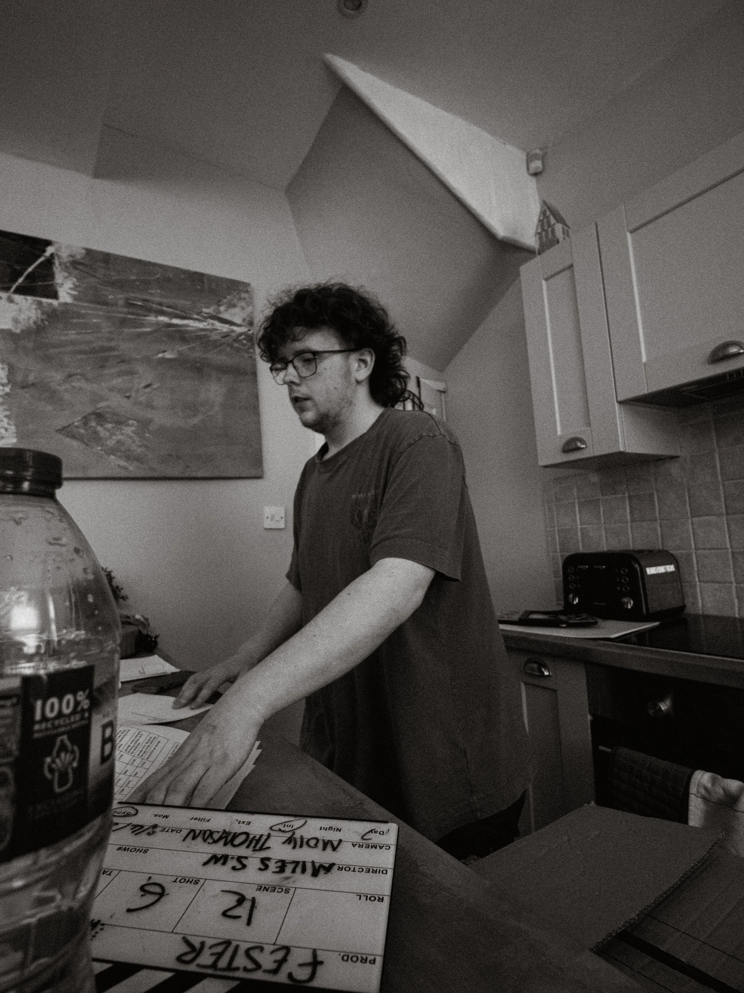 A young man with curly hair and glasses, wearing a T-shirt, standing in a kitchen, writing or working on a piece of paper on the table. The kitchen has white cabinets and a toaster on the counter. There is a painting on the wall and a bottle of water on the table.