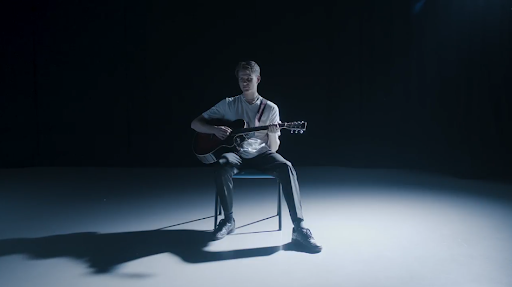 A person sitting on a chair playing an acoustic guitar on a dark stage.