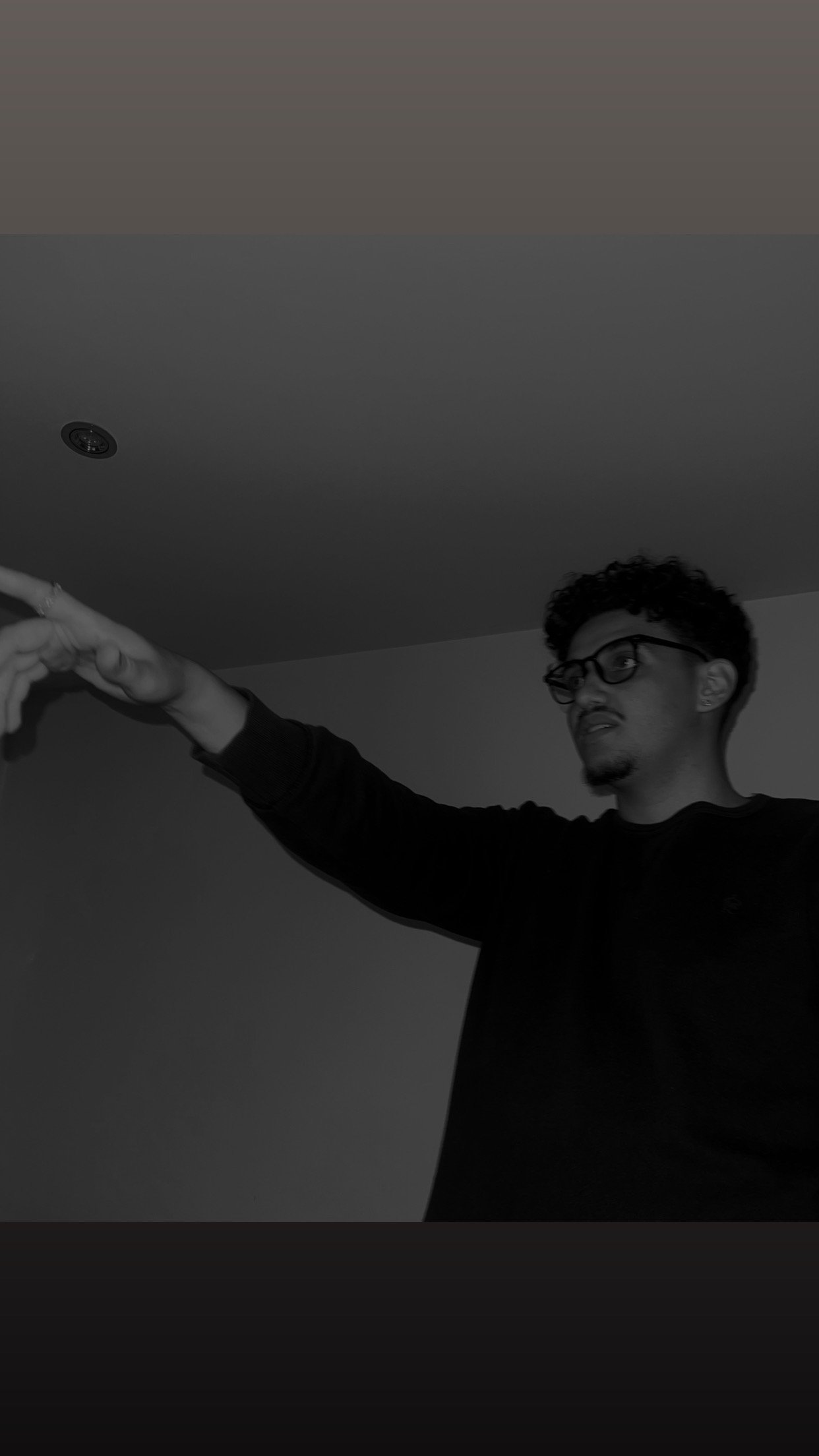 A young man with glasses and curly hair gesturing with his right hand in a room with plain walls and a ceiling light.