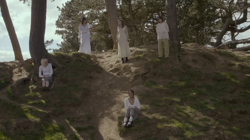 Five women in white and beige clothing outdoors among trees, some standing on a hill and one sitting on the ground.