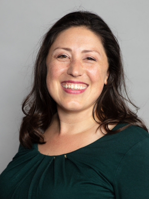 A woman with dark brown hair smiling against a plain gray background.