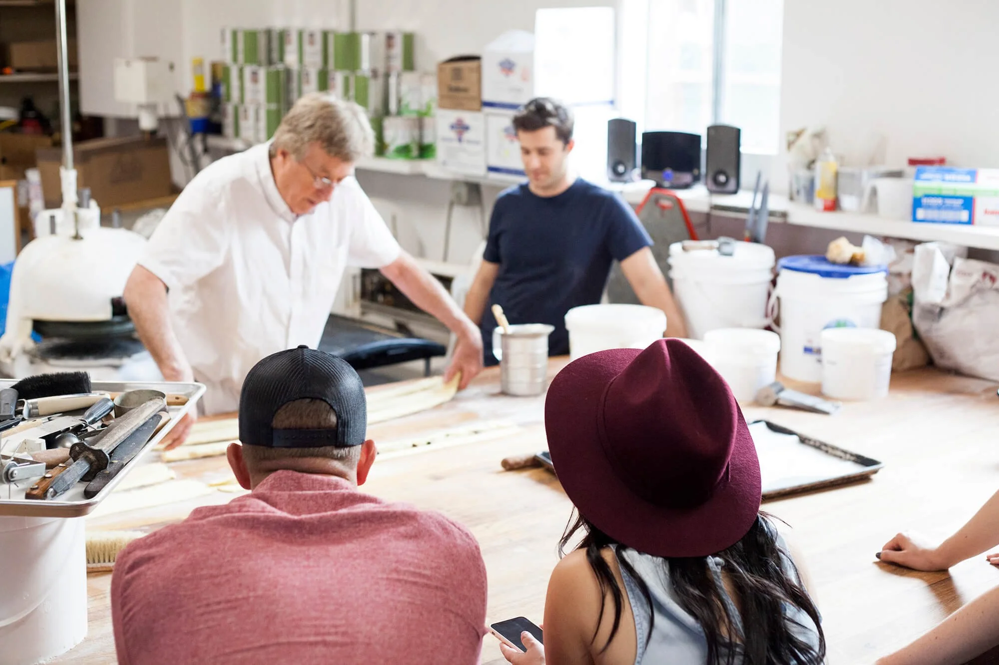 A group of people gathered around a workshop table where two men are demonstrating woodworking, while two women watch