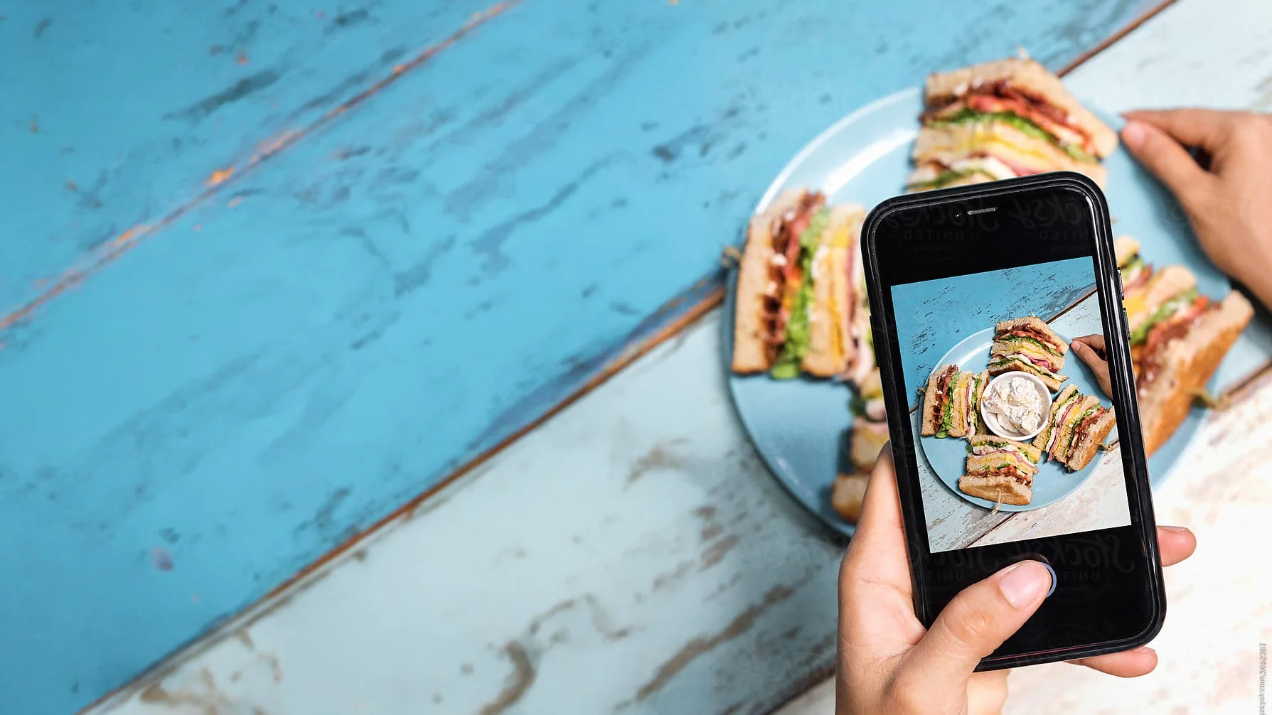 Person taking a photo of a plate of sandwiches and a bowl of dip on a distressed blue wooden table with a smartphone.