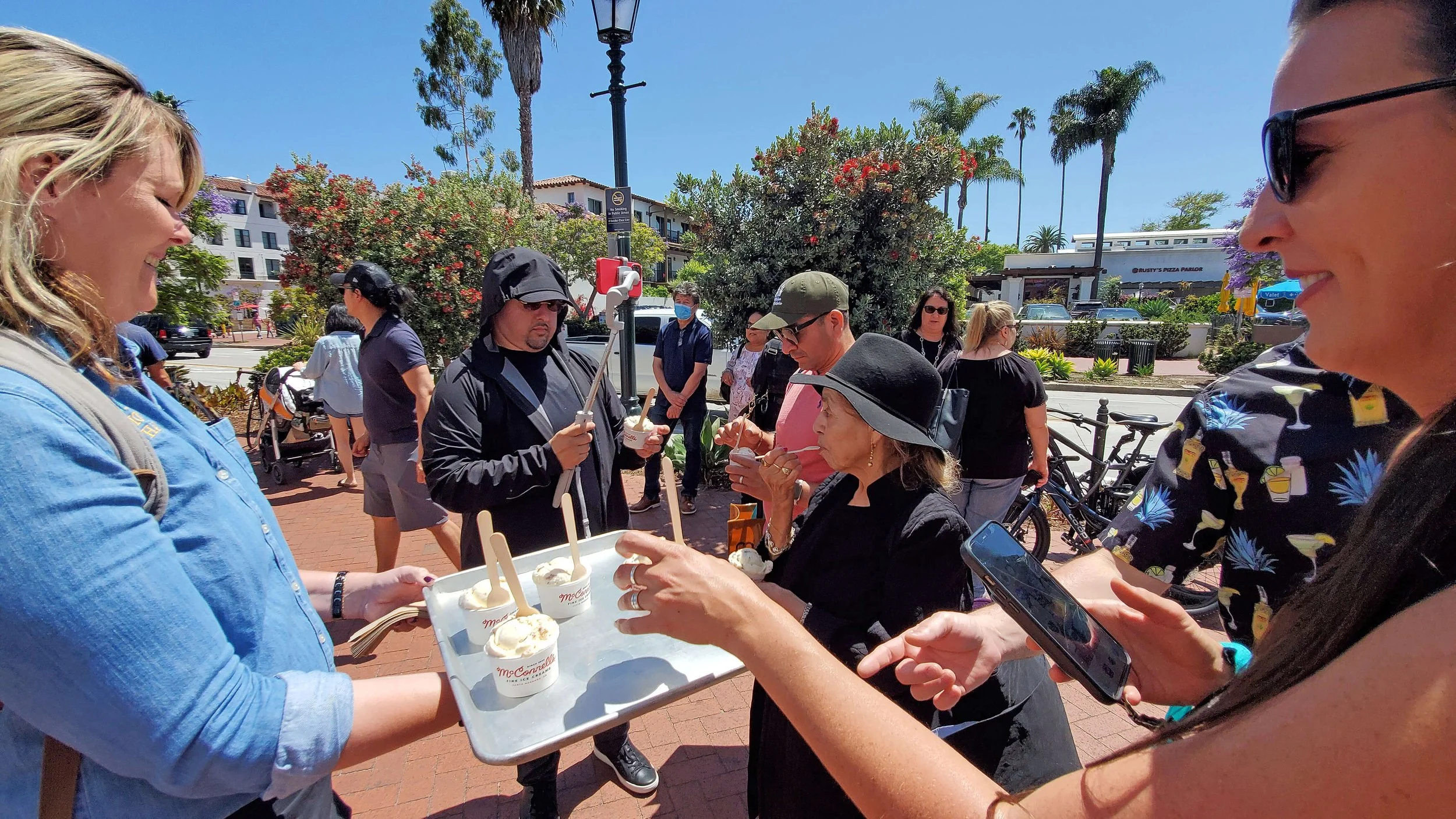 Group of people outdoors on a sunny day, some eating ice cream, others waiting or talking, with trees and buildings in the background.
