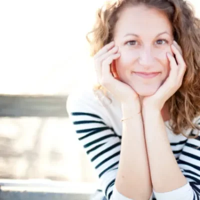 A woman with curly hair smiling, resting her face in her hands, wearing a striped shirt, outdoors with a blurred background.