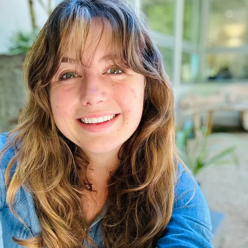 Close-up of a smiling woman with long wavy brown hair, wearing a blue shirt, indoors with a blurred background of windows and plants.