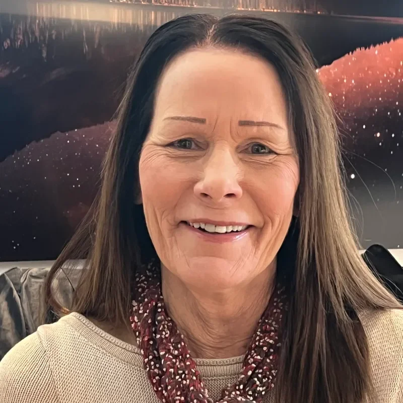 A woman with long brunette hair smiling in front of a backdrop resembling a starry night sky over a body of water during sunset or sunrise.