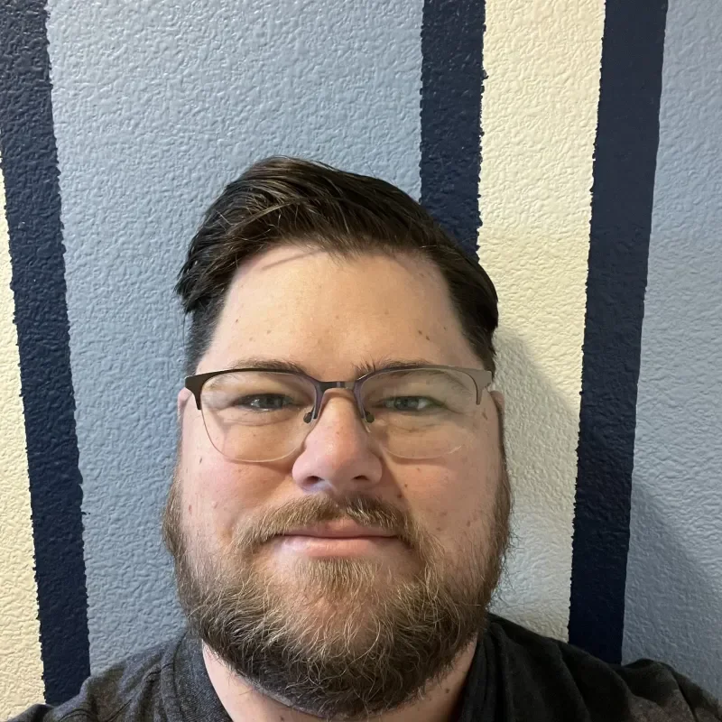 A young man with glasses and a beard taking a selfie against a textured wall with vertical navy blue and beige stripes.