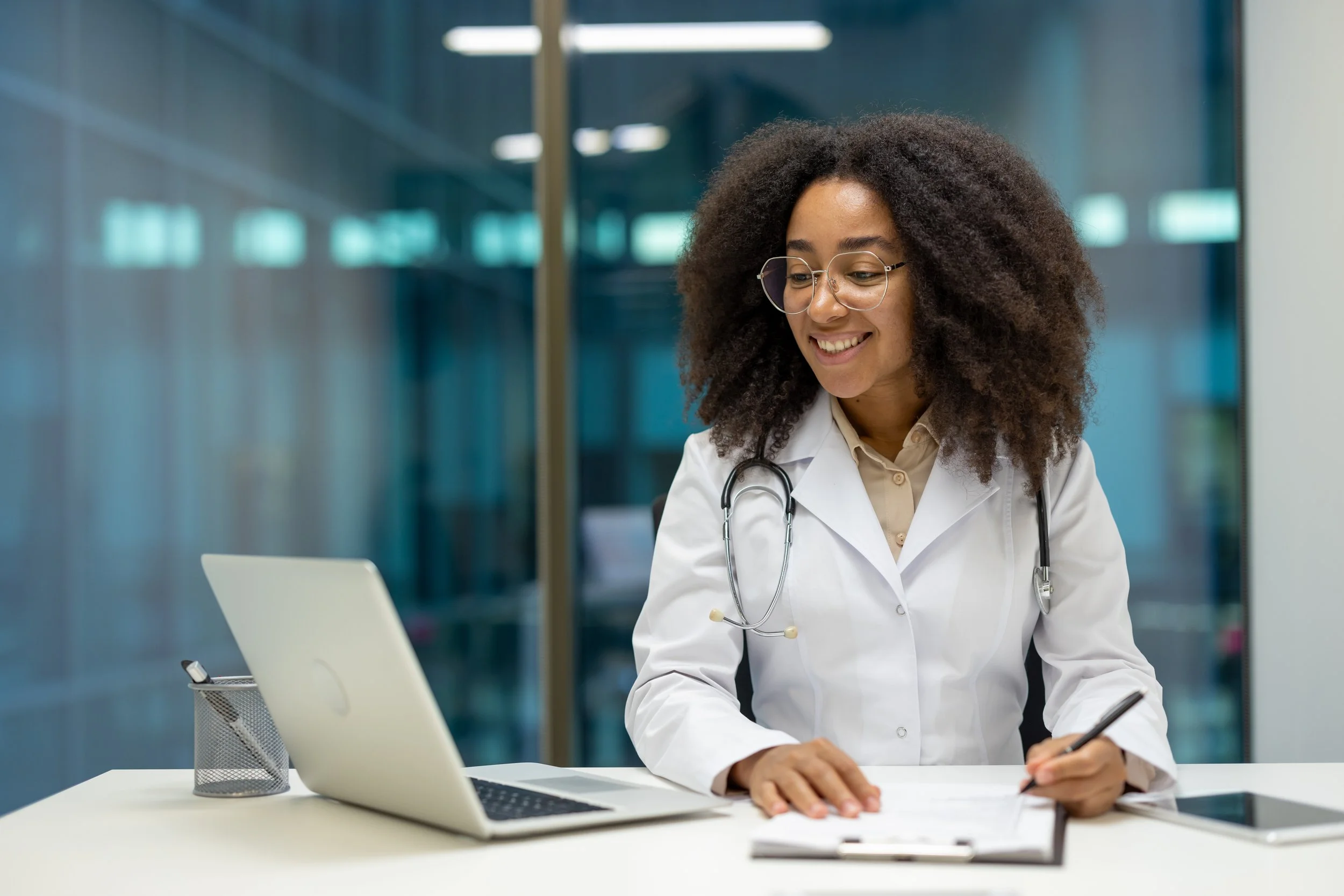 A female doctor with glasses, curly hair, and a stethoscope around her neck sitting at a desk, smiling while writing on a notepad with a laptop and a tablet nearby.