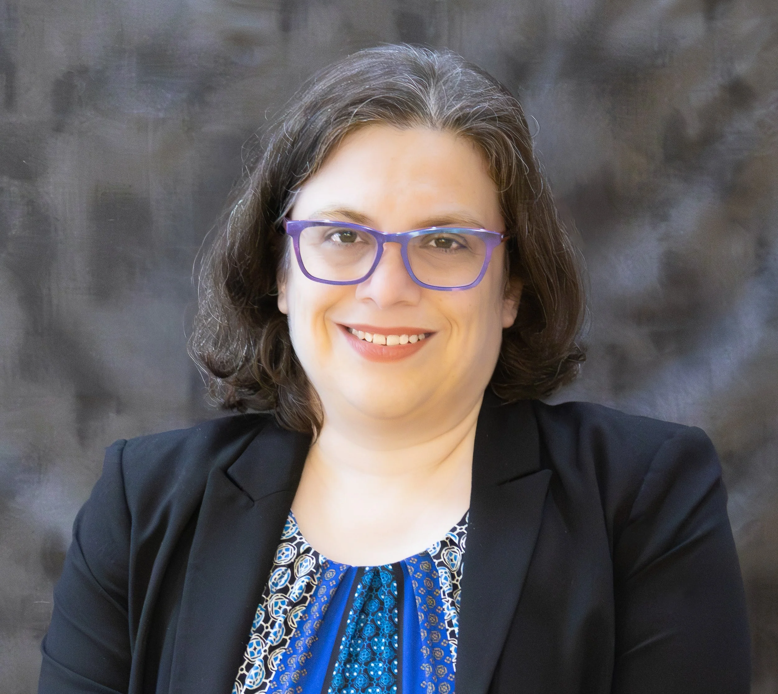 A woman with shoulder-length brown hair, glasses, and a black blazer smiling at the camera against a dark, textured background.