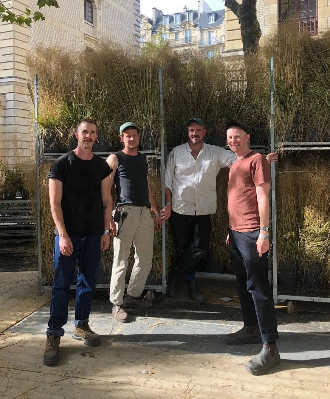 Four men standing outdoors in front of tall grass and trees, smiling at the camera. They are dressed casually and appear to be in an urban park or garden setting.