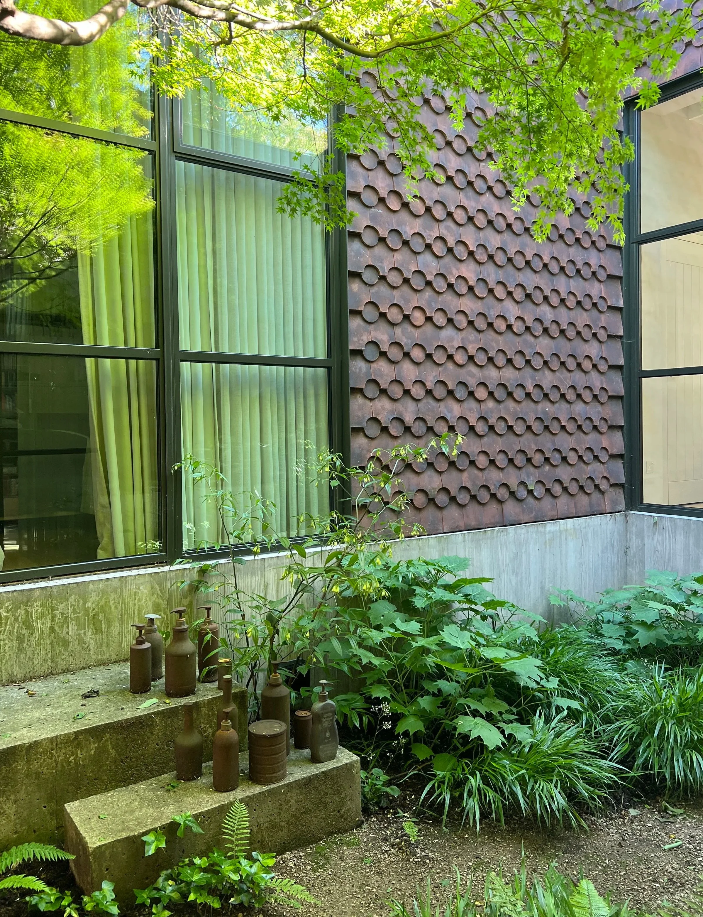 Green foliage and trees surround the exterior of a modern building with large windows and a decorative brown wall with circular tiles. Several small ceramic bottles are placed on concrete steps near lush green plants.
