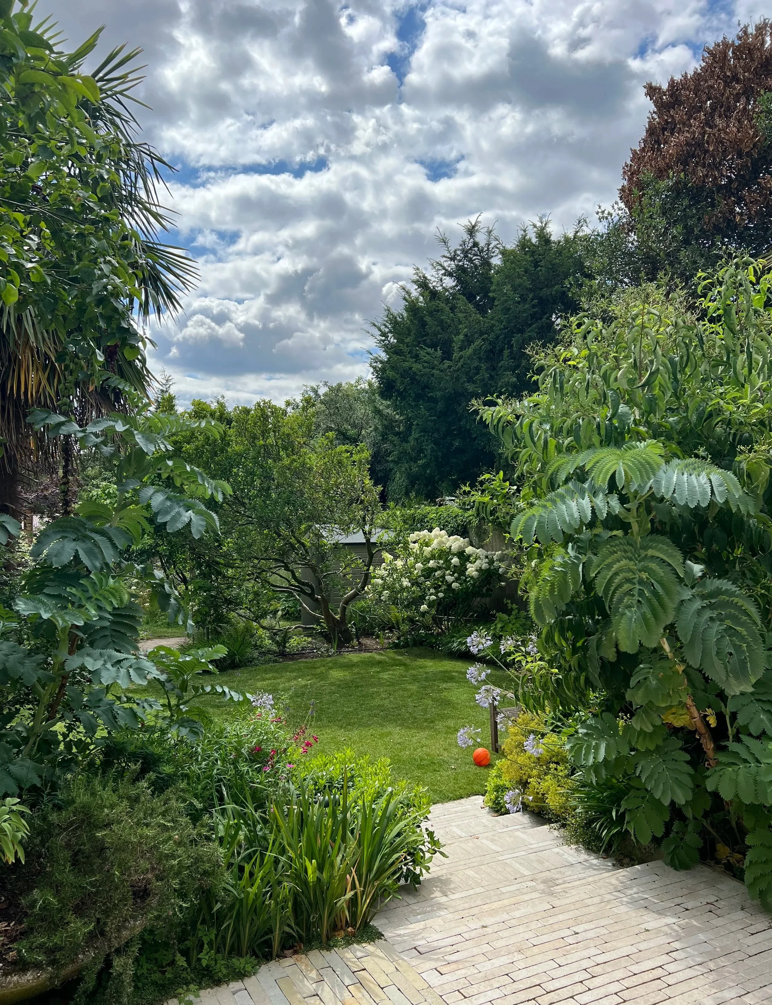 A lush green garden with various flowering plants, bushes, and trees. There is a small, well-kept lawn area, and the sky above is partly cloudy with patches of blue.