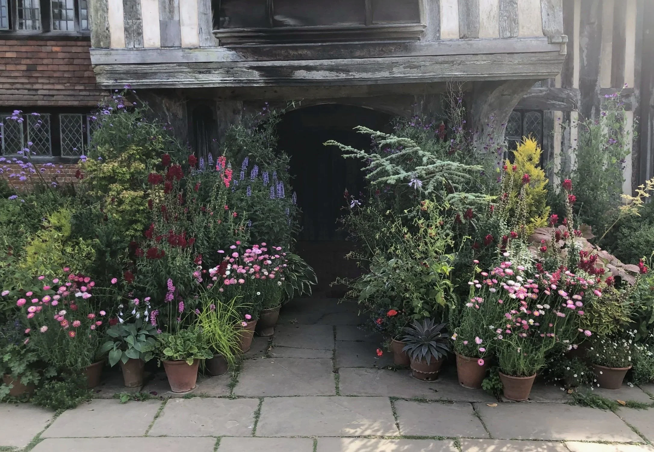 A front view of a house entrance with an archway surrounded by potted and planted colorful flowers and greenery, with stone paving in the foreground.