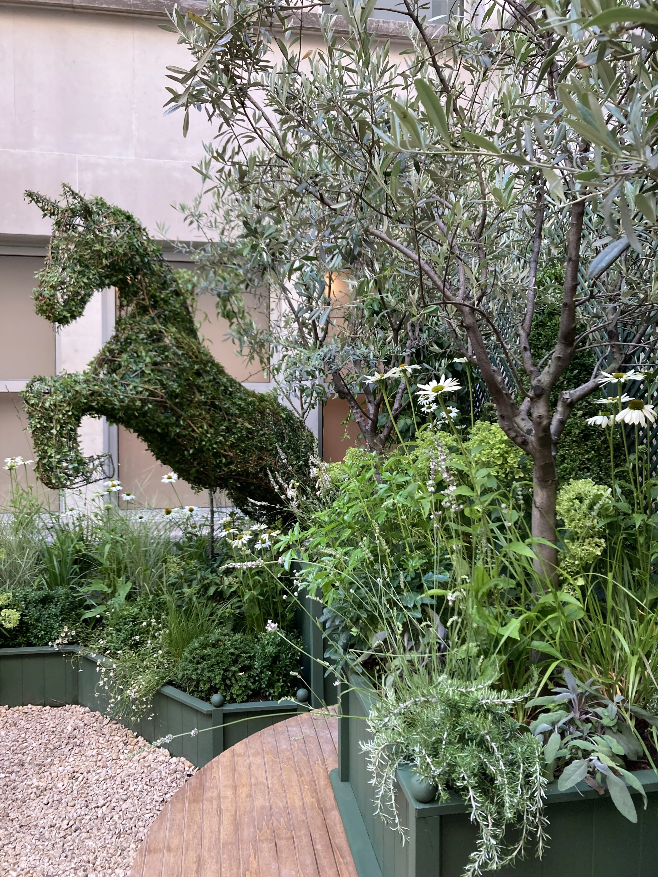 A garden with various green plants, flowers, and trees, including a topiary shaped like an animal, next to a small wooden pathway.