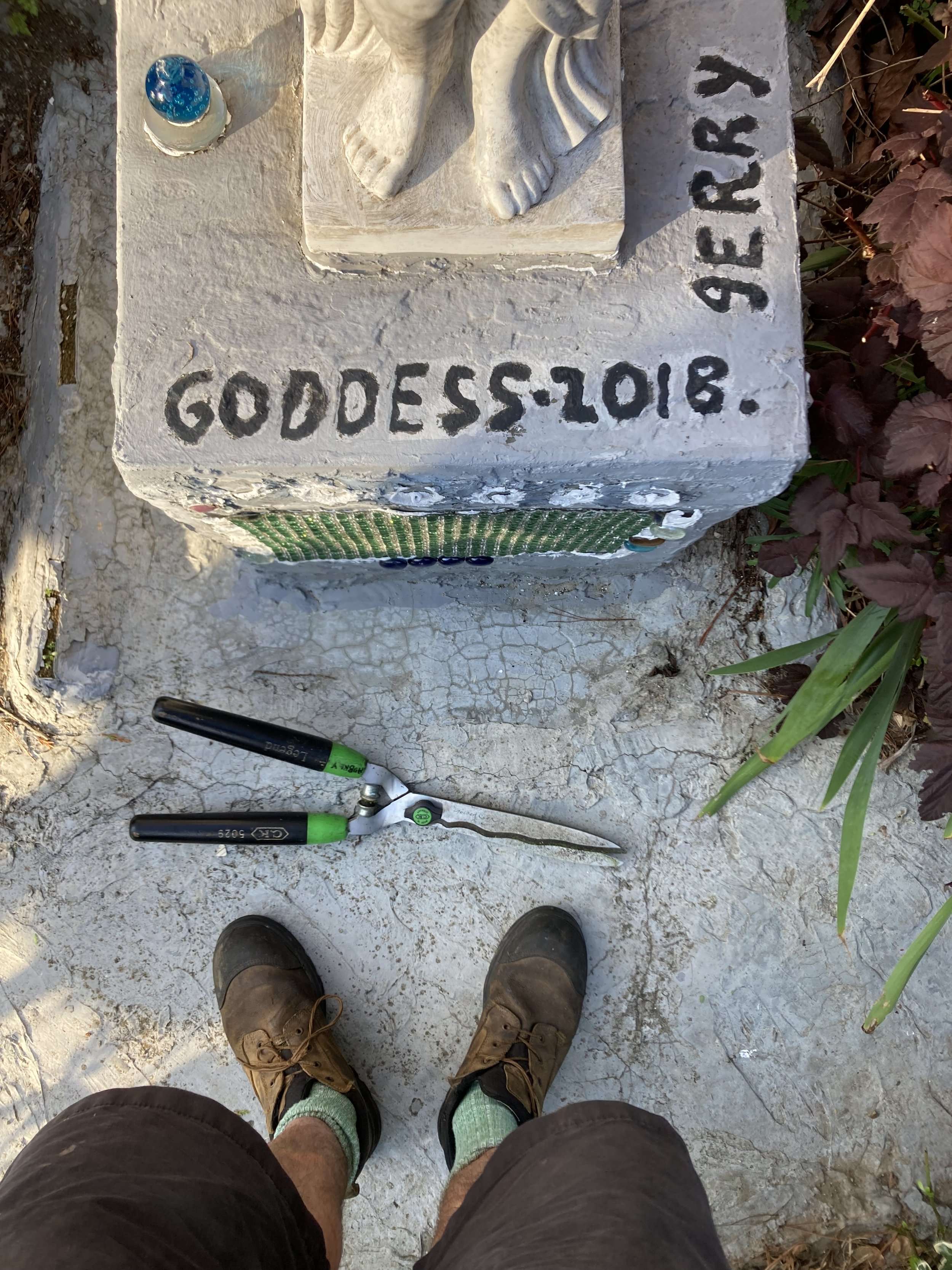 A concrete monument features the inscription 'GODDESS-2018' and 'GIVER.' There is a small blue decorative object, a pair of hedge shears, and a person's feet in brown shoes with green socks, standing on a cracked concrete surface with plants nearby.