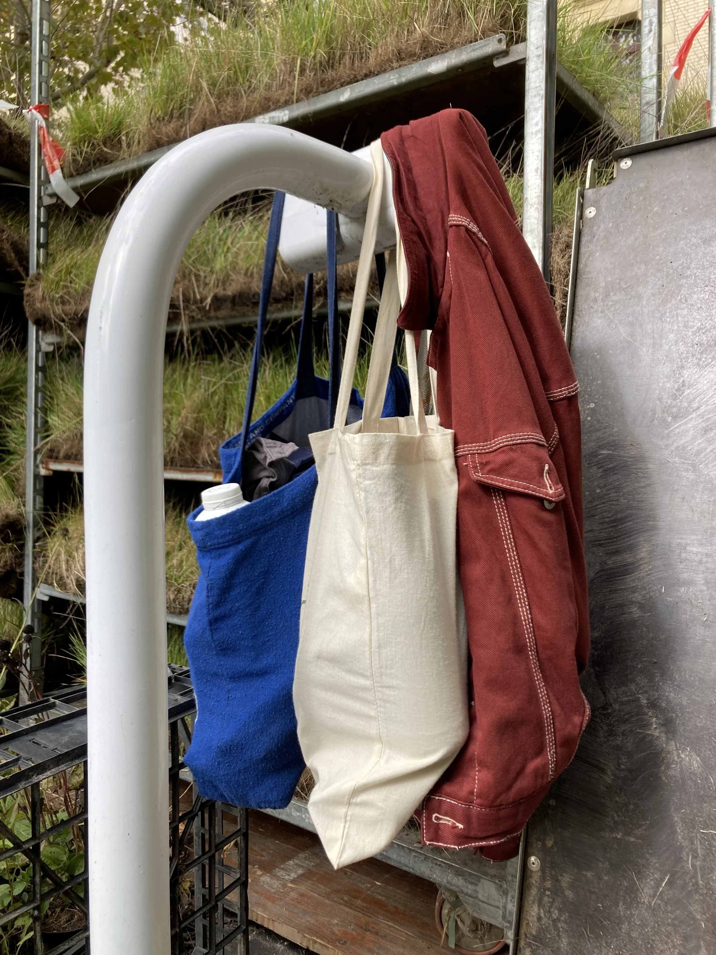 Hanging bags and a red jacket on a white metal bike rack in an outdoor garden or nursery area.