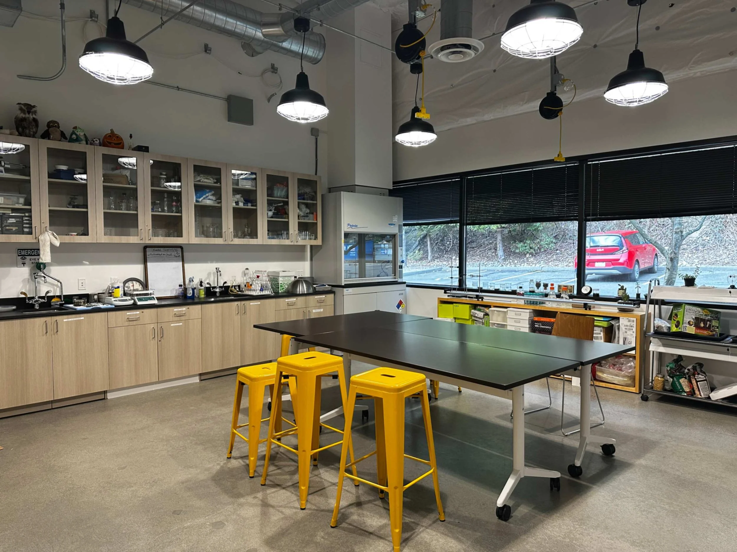 The science classroom of �ǿ����޴�ý with wooden cabinets filled with glassware and supplies, a laboratory table with yellow stools, and a large window with black blinds showing an outdoor view.