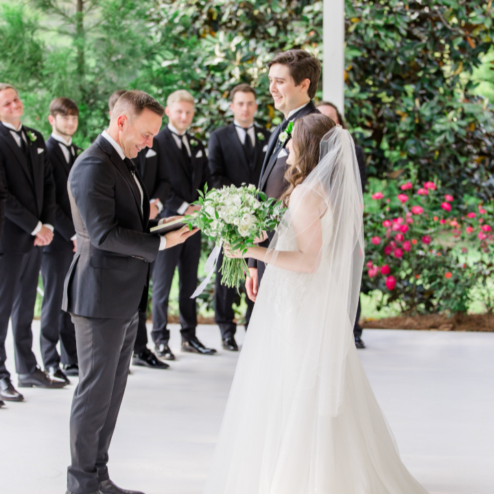 Bride and groom exchanging vows during a wedding ceremony with groomsmen in black suits standing behind them and lush green foliage in the background.