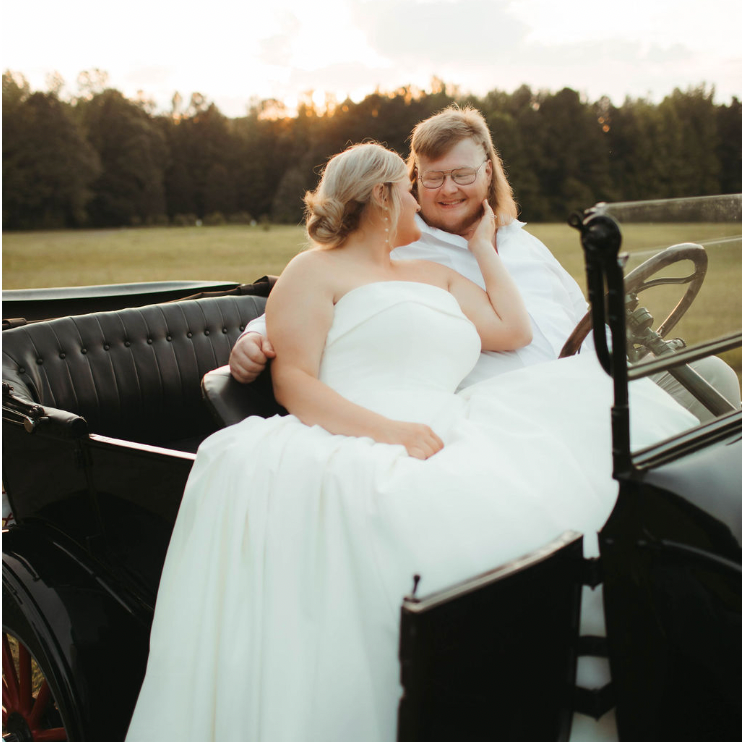 Couple in wedding attire sitting in a vintage car, embracing outdoors at sunset, with a field and trees in the background.